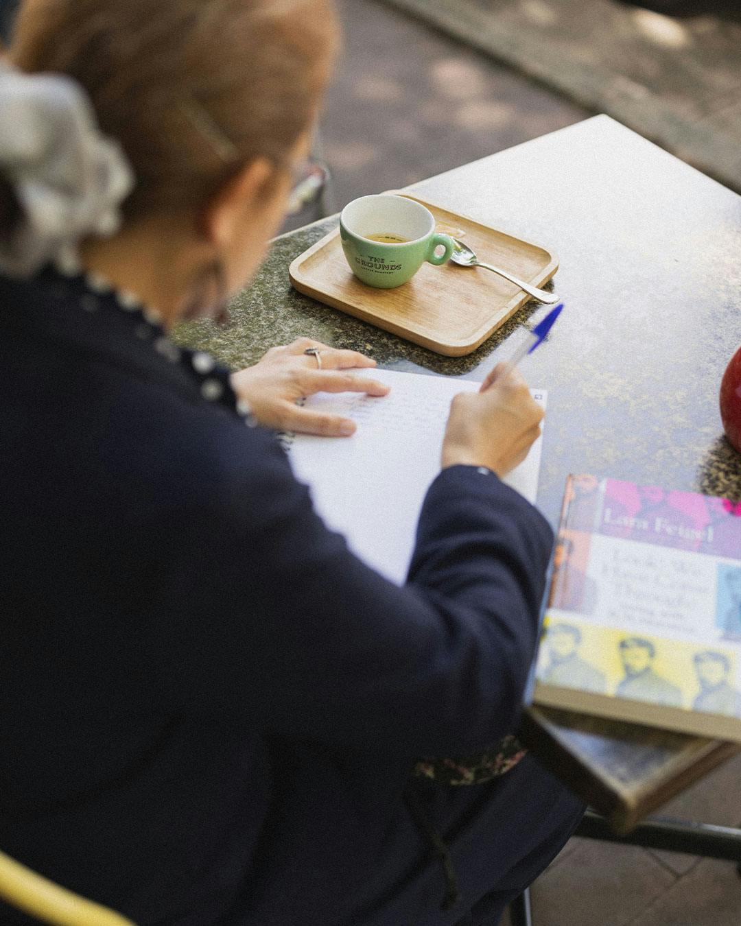 Yumna Kassab writing in a notebook at a table with a book and a tray holding a coffee cup.