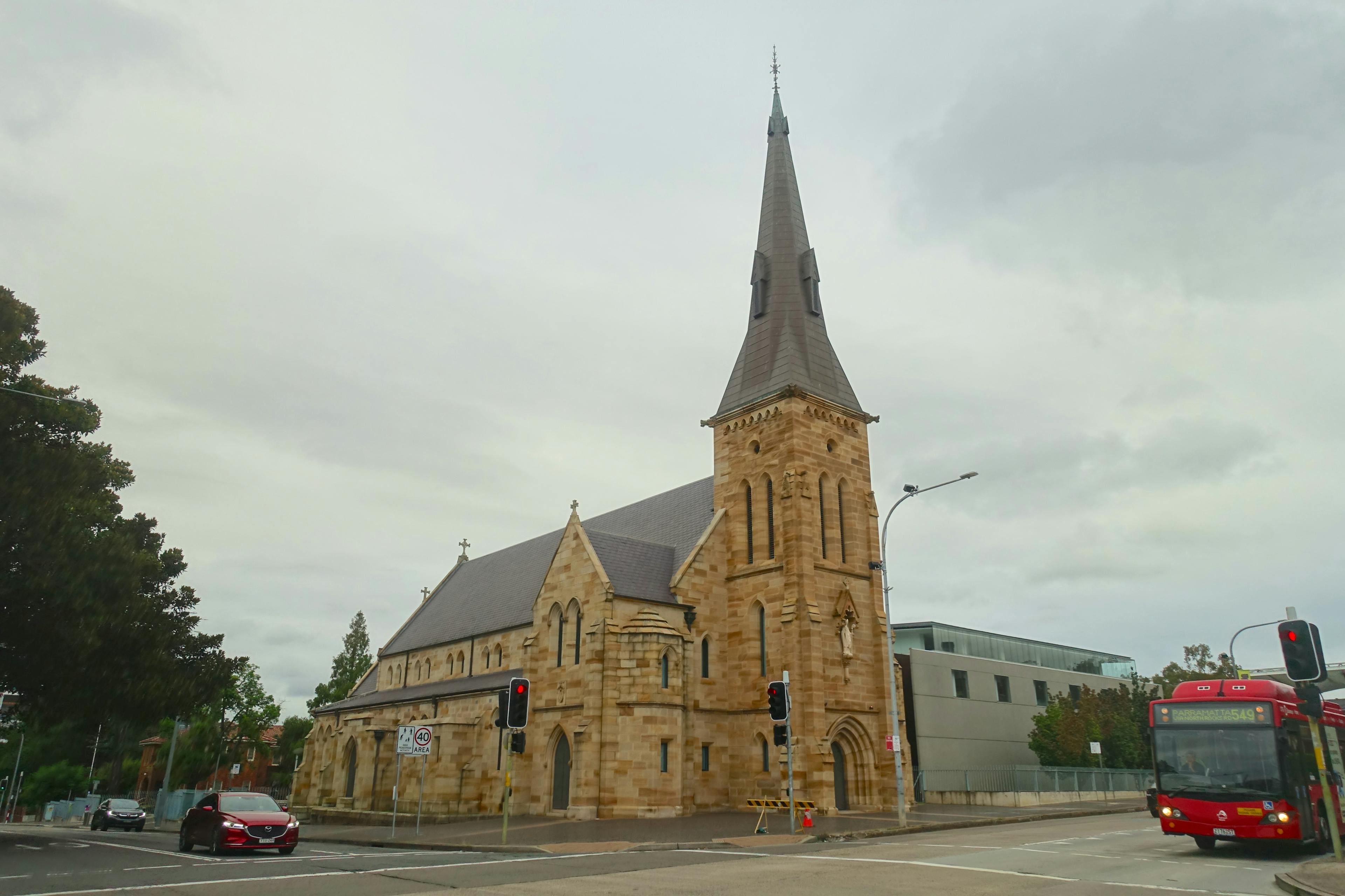 Street corners of Victoria Road and Marist Place in Parramatta, with a church and tall tower set further back. 