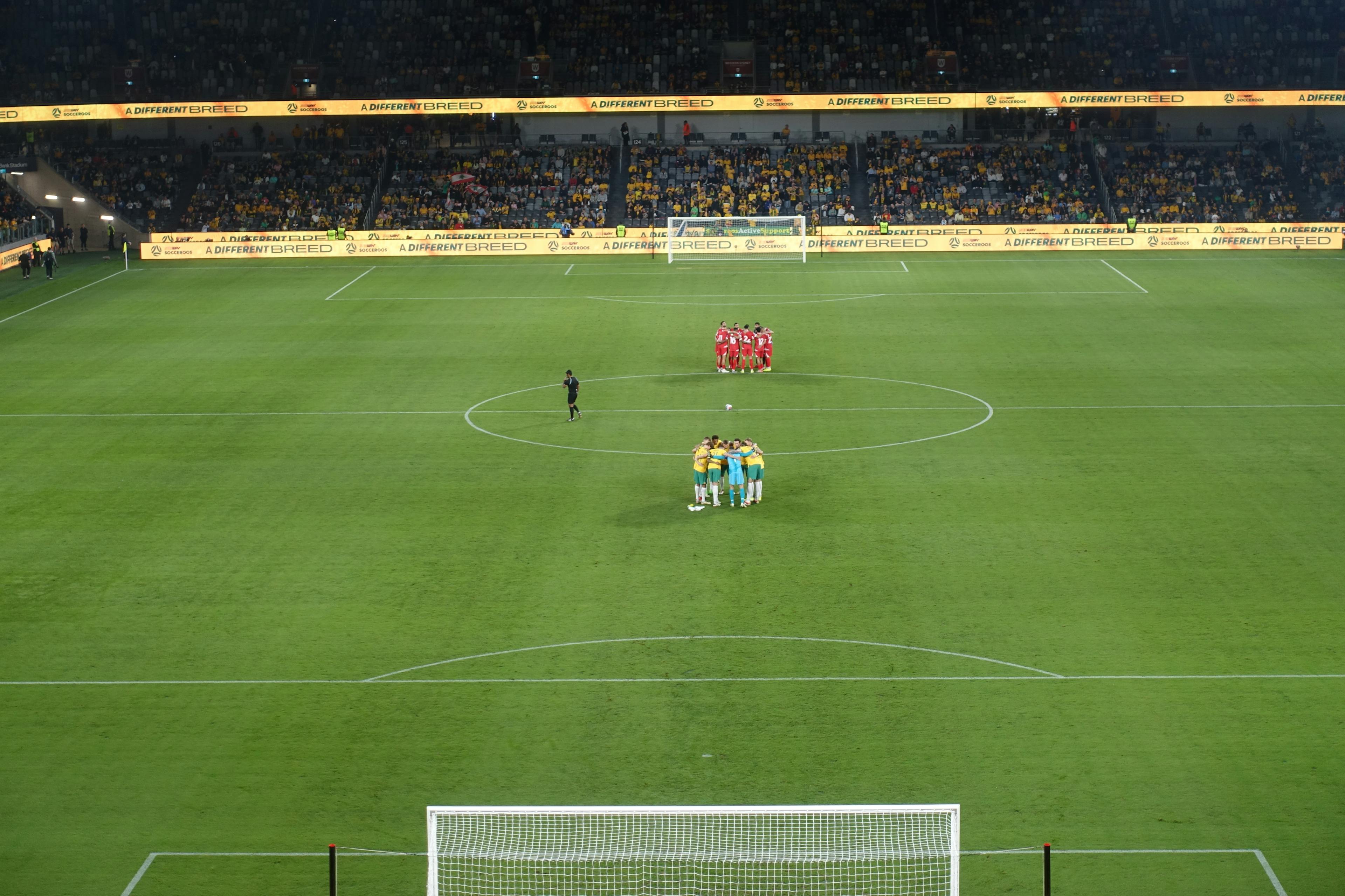Two soccer teams in separate huddles on the field before a match at Parramatta Stadium. 