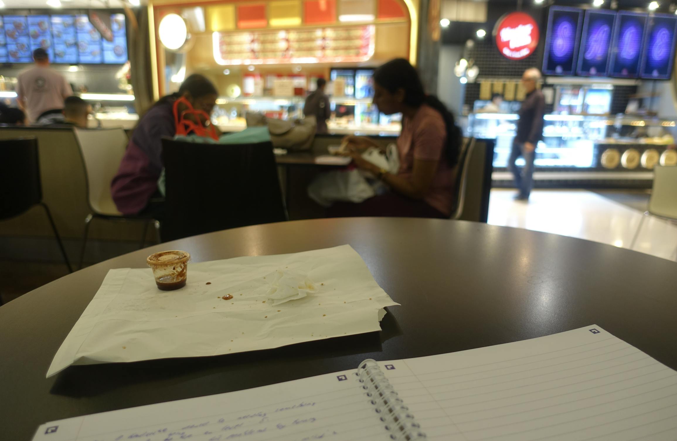 A notebook with writing, an empty paper bag, and a sauce container on a table at the Parramatta Westfield food court, with blurred people and stalls in the background.