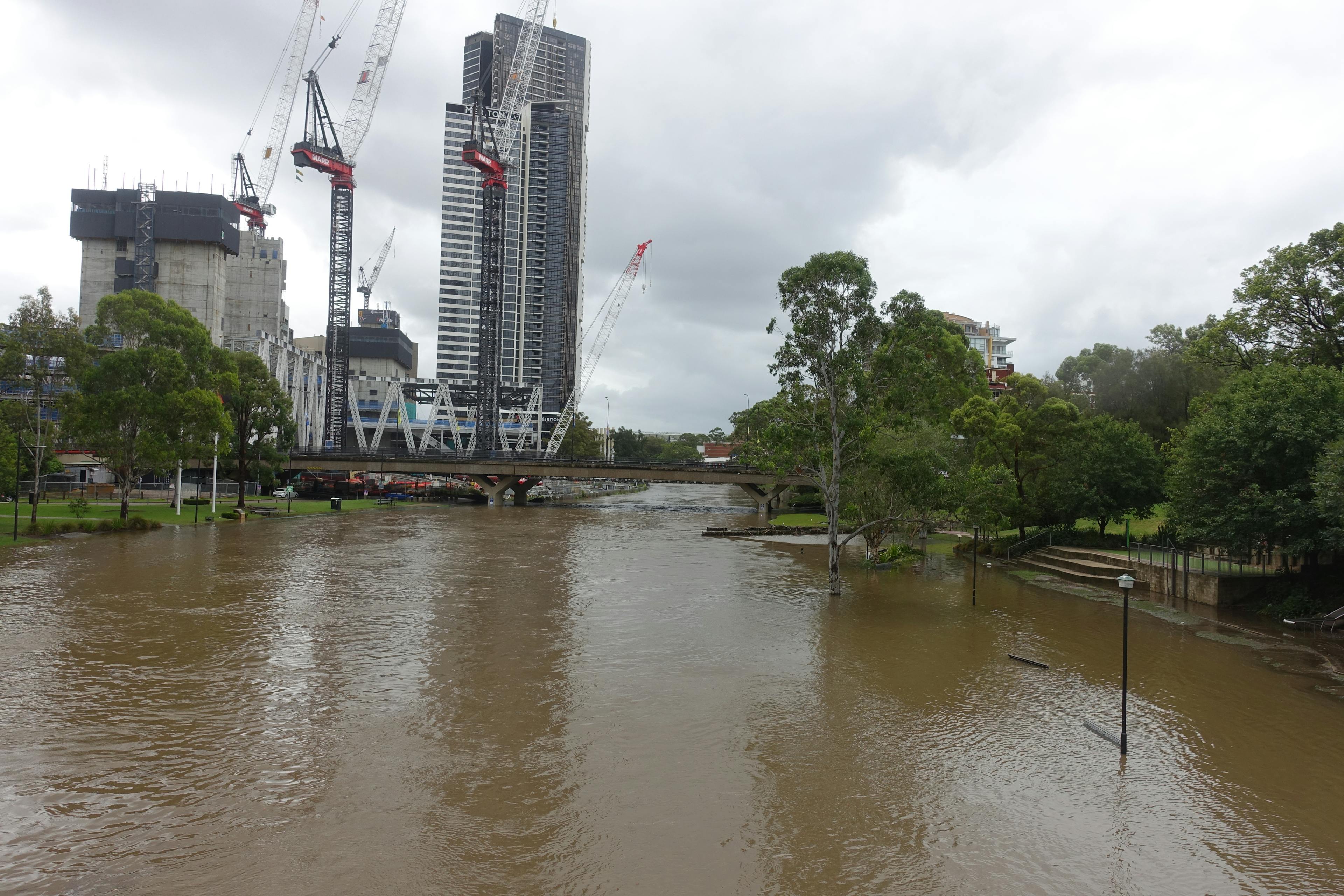 Flooded river foreshore with a submerged bin and two pedestrian bridges nearly underwater; no benches visible. 