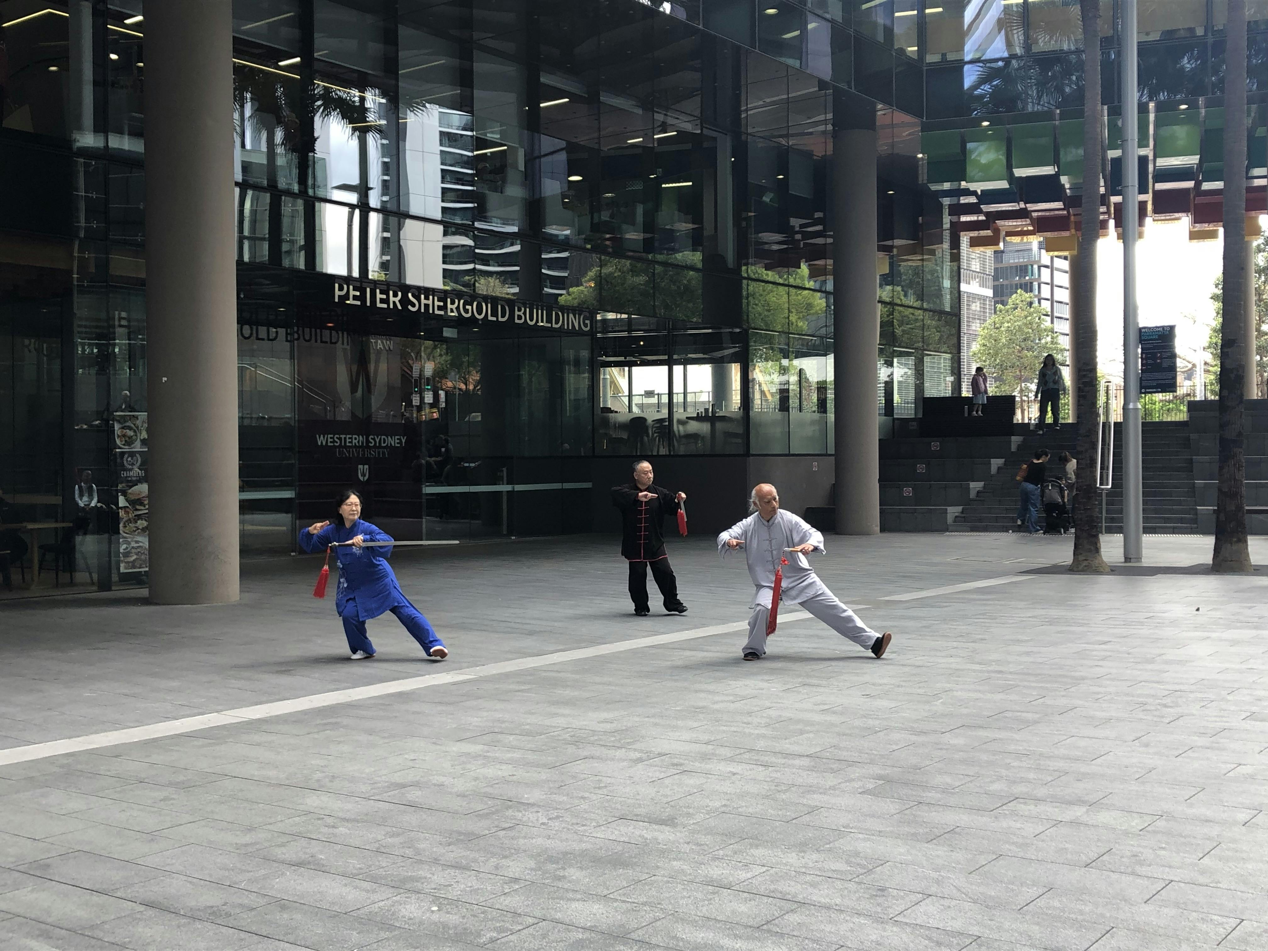 Three people practicing Tai Chi in Parramatta Square, in front of the Peter Shergold Building.  