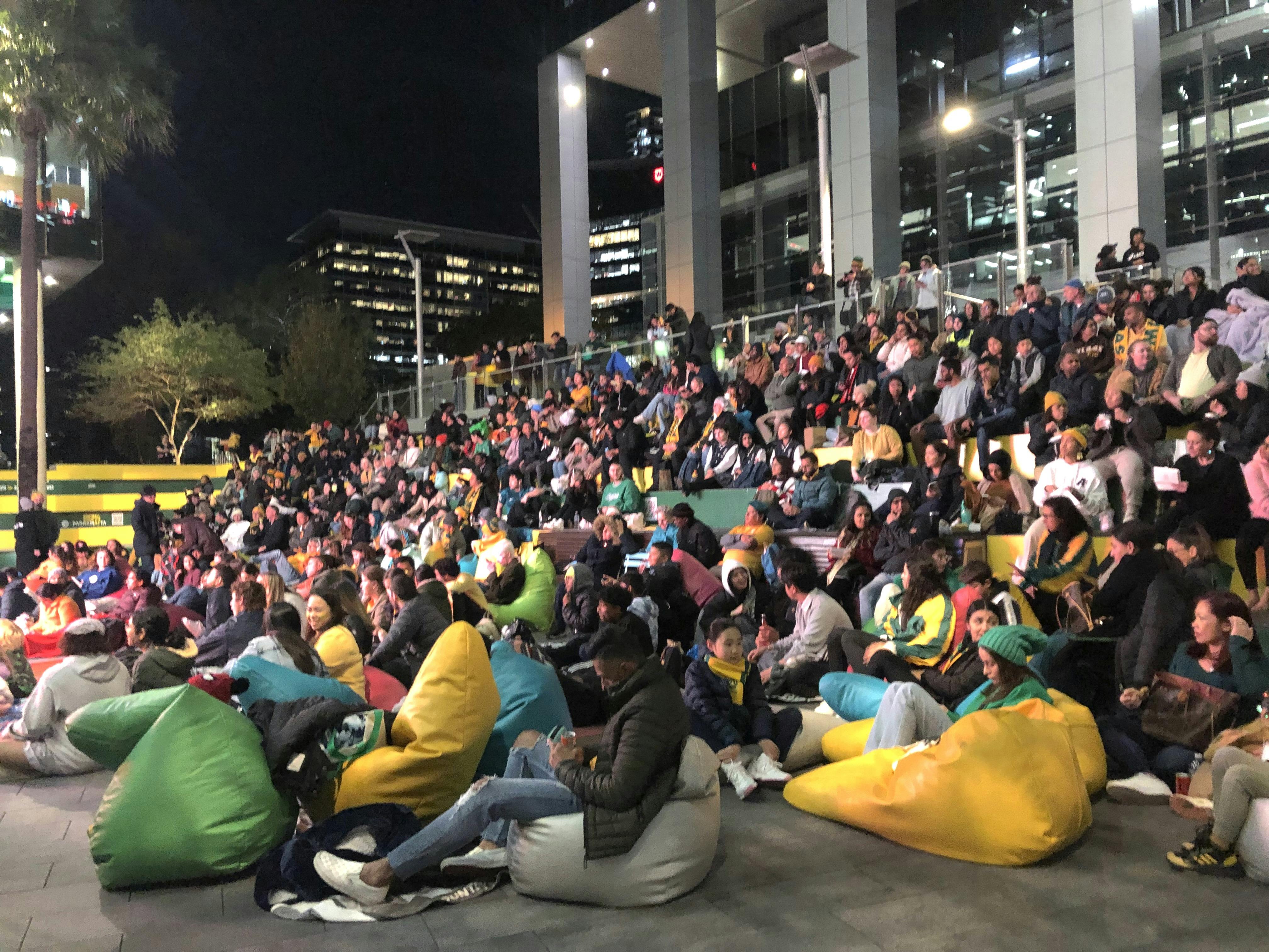 A crowd of people sitting on bean bags and the steps in Parramatta Square, watching a large screen.  