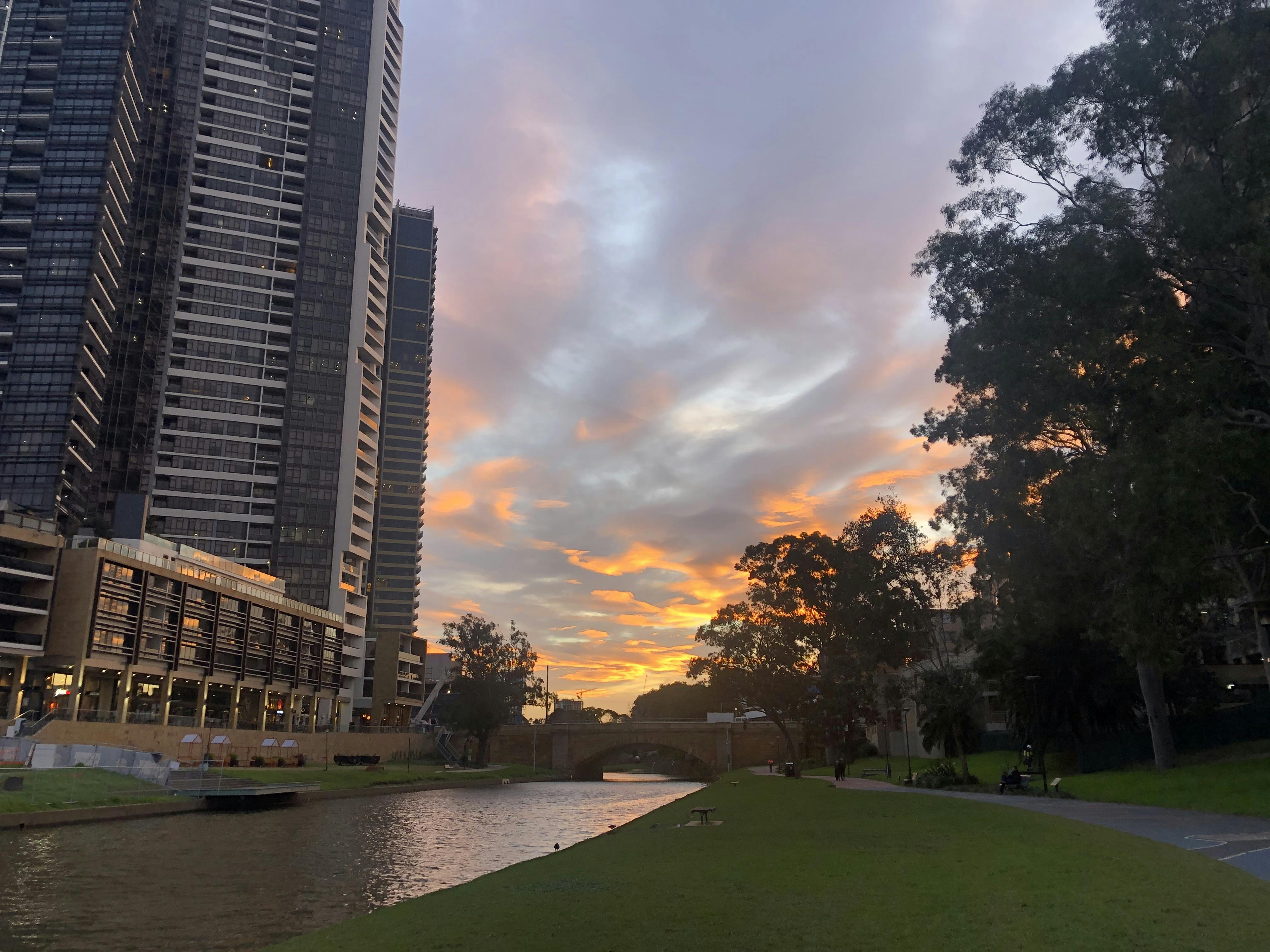 Photo of Parramatta River from Parramatta Park at sunset. A wide grass area and walking path are on the right side, tall buildings are on the left, a bridge is visible in the distance, and the sky blends orange, white, blue, and grey.
