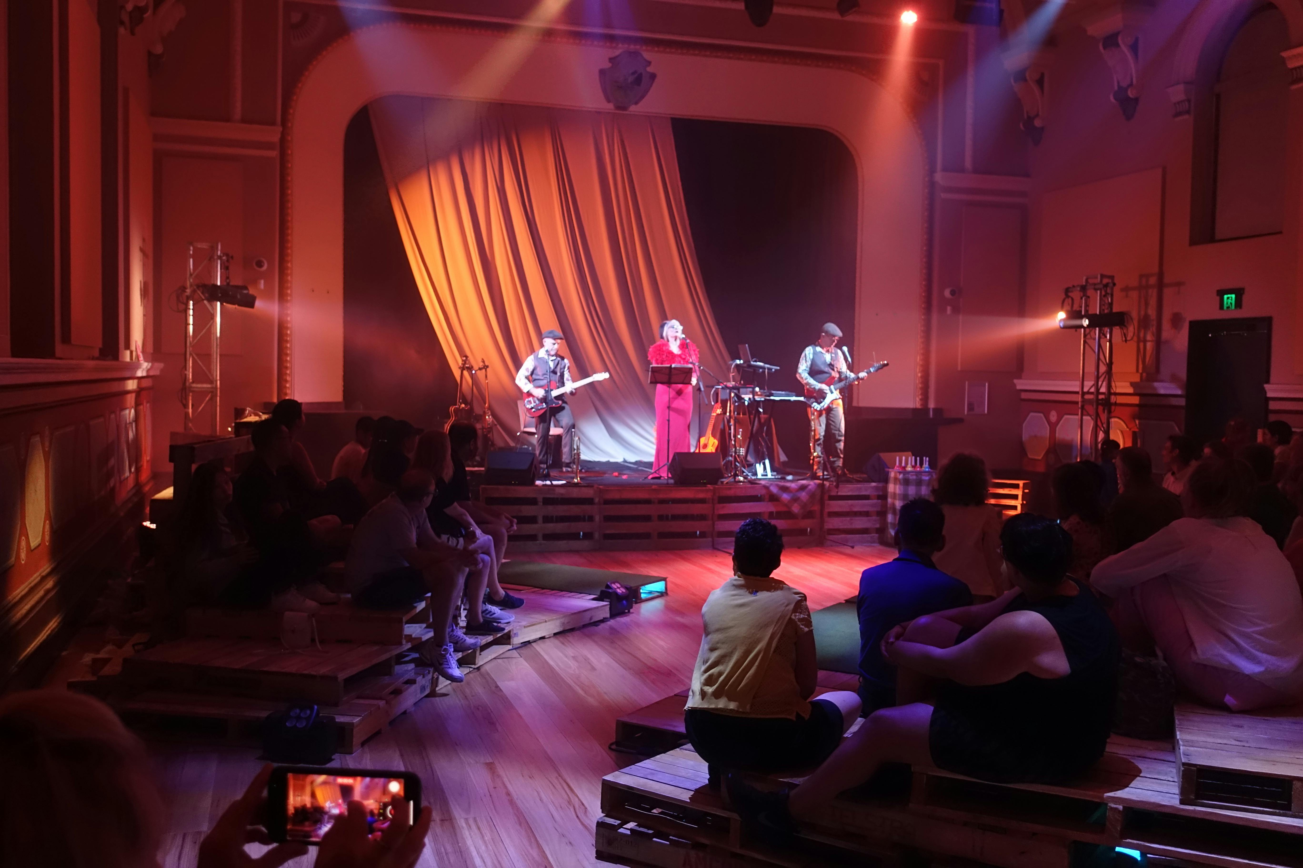 A band performing on stage to an audience seated on the floor and wooden steps. 