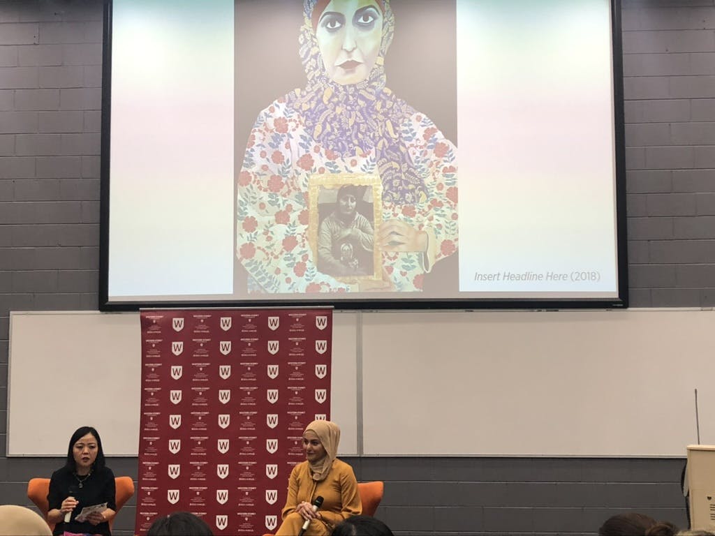 The artist Amani Haydar sitting on a stage with an interviewer. There is a large screen behind her with a photo of her painting ‘Insert Headline Here’.