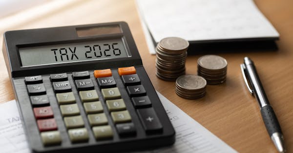 A close-up of a calculator displaying “TAX 2026” on a wooden desk, with stacks of coins, a pen, and a notebook in the background, suggesting financial planning or tax preparation.