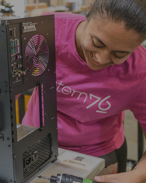 Woman in pink System76 tshirt holds a screw and smiles as she builds a Thelio desktop.