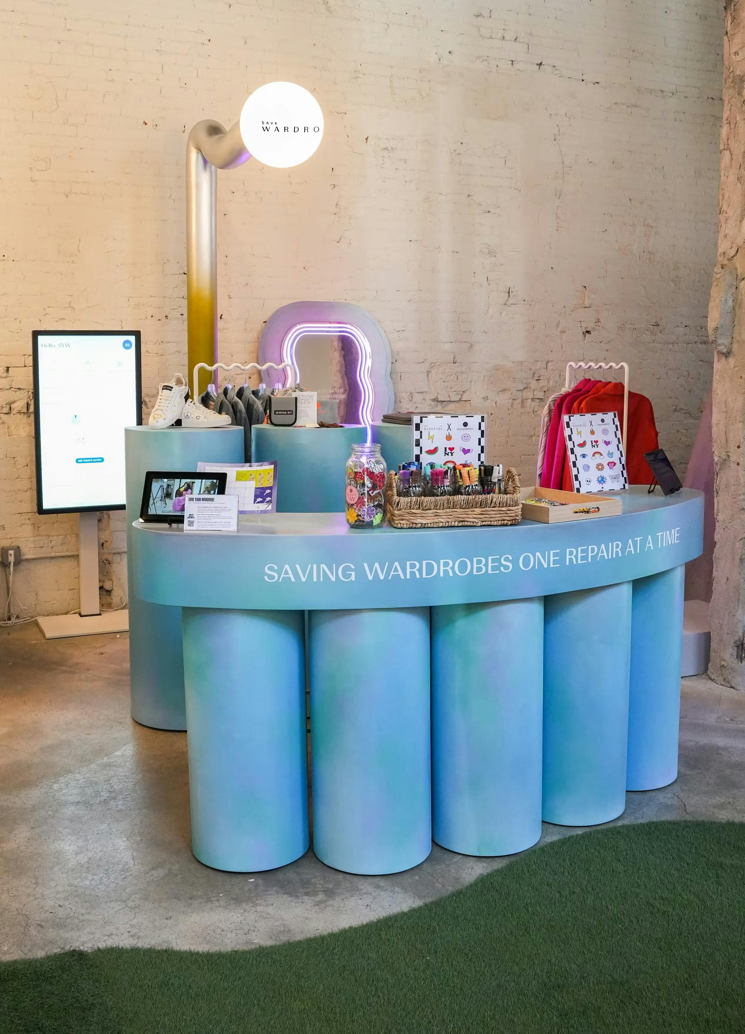 The Save Your Wardrobe pop-up space at Showfields Noho, image shows blue 'Repair Bar' which reads 'Saving Wardrobes one Repair at a Time'  with sewing utensils on top. The space also features a digital wardrobe activation and upcycled garments on clothes rails.