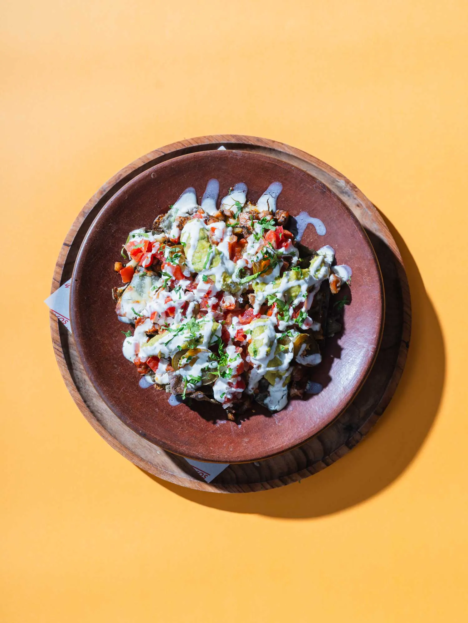 Mexican nachos topped with seasoned meat, melted cheese, guacamole, pico de gallo, sour cream drizzle, and fresh cilantro, served on a rustic clay plate at a Mexican restaurant in Umalas Bali