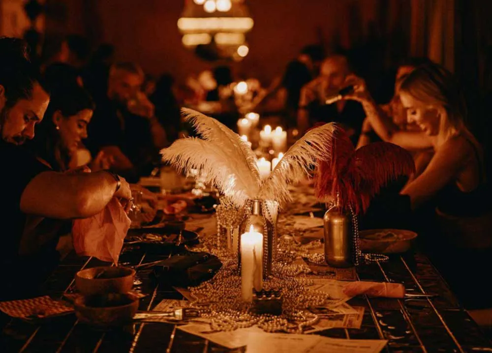 Elegant candlelit dinner party with gold fringe chandelier, white ostrich feather centerpieces, and guests seated at a long decorated table