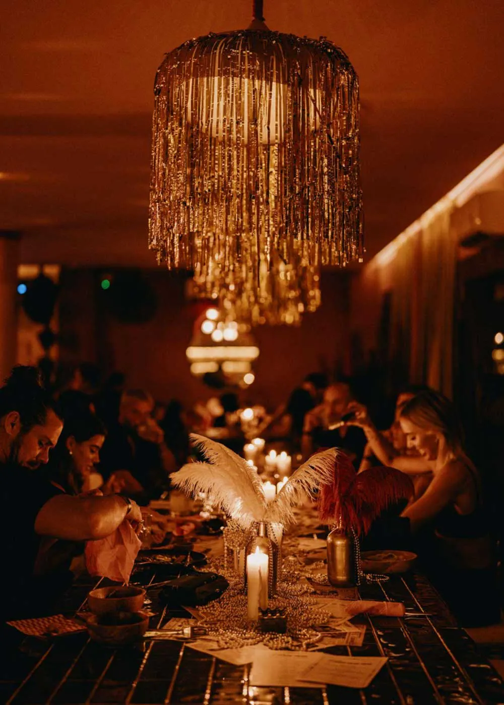 Elegant candlelit dinner party with gold fringe chandelier, white ostrich feather centerpieces, and guests seated at a long decorated table