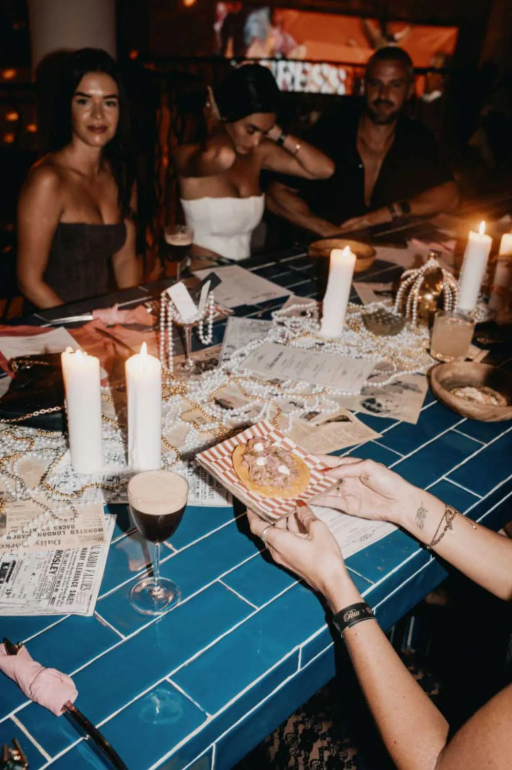 Group of people enjoying tacos and cocktails at a candlelit table decorated with pearls and newspapers, creating a festive and intimate dining atmosphere in Uluwatu, Bali