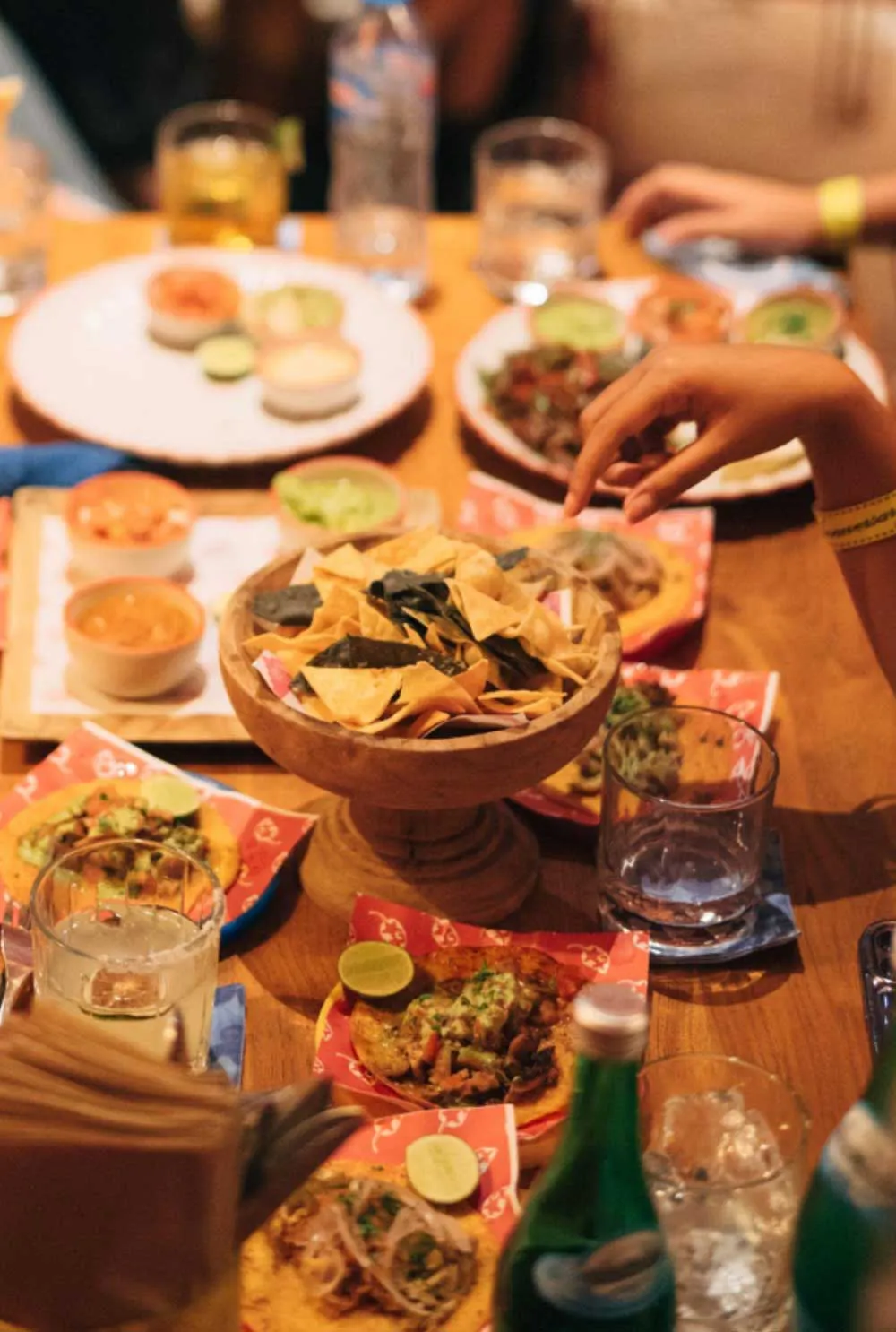 Wooden table with a bowl of tortilla chips surrounded by small tacos topped with lime, guacamole, and salsa, ready to serve at a casual Mexican dining experience in Bali
