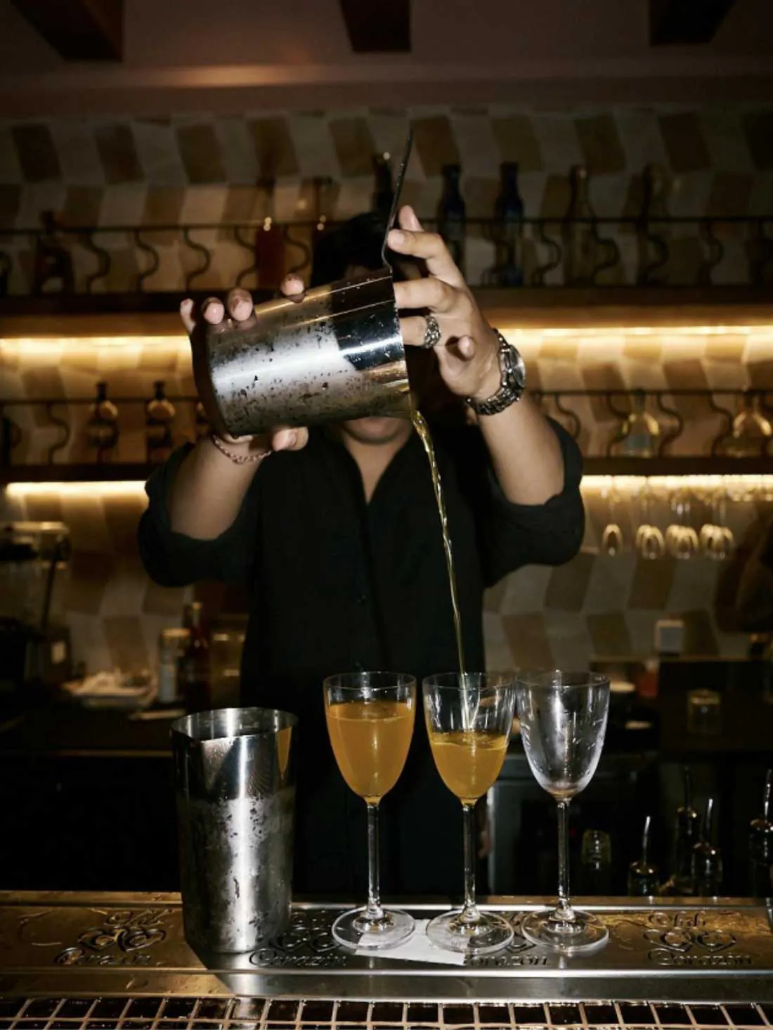 Bartender pouring cocktails from a metal shaker into three stemmed glasses at a stylish bar with warm lighting and shelves of spirits in the background.