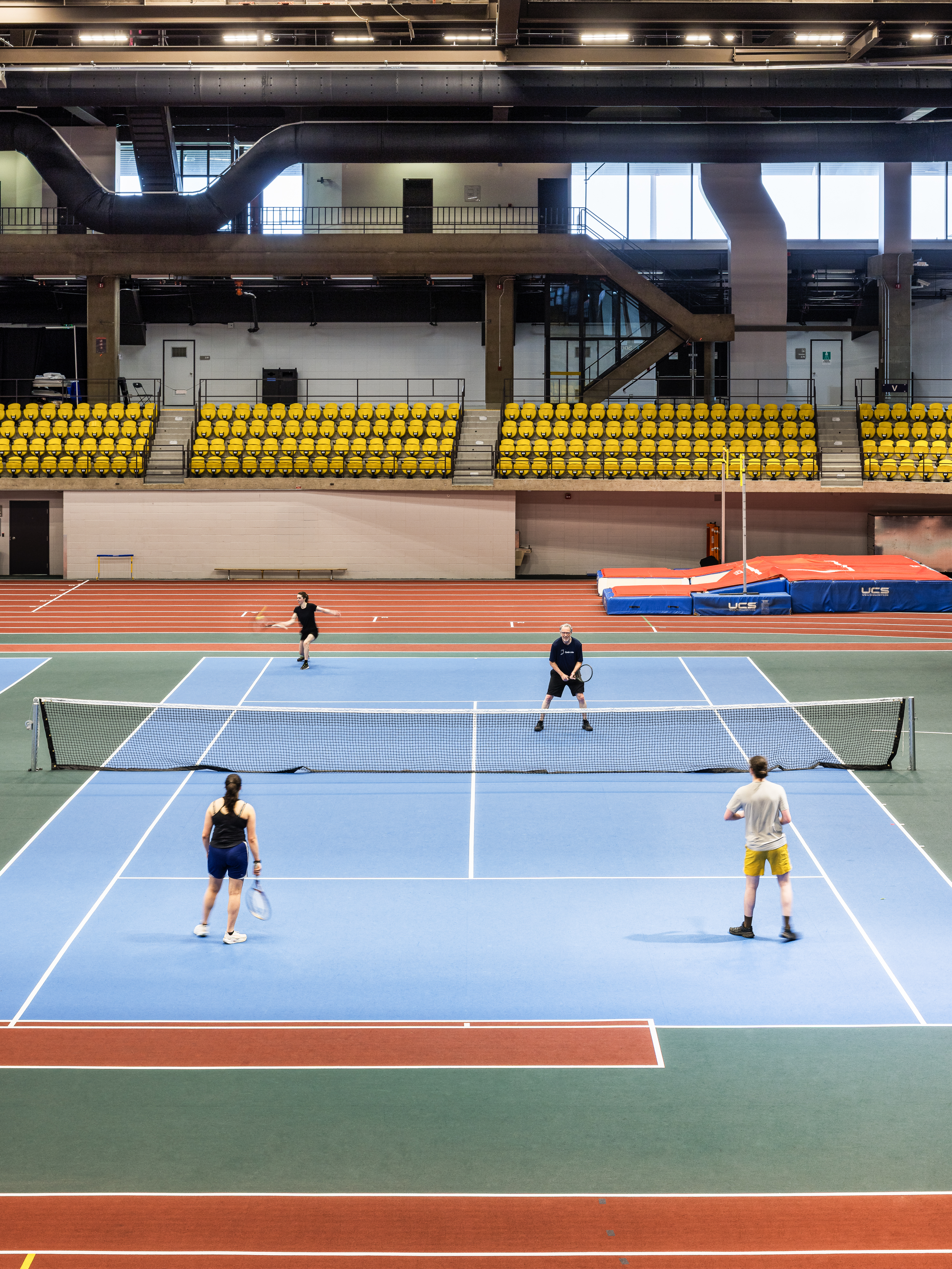 Architectural and design photography, players on an indoor tennis court, yellow bleachers in the background.
