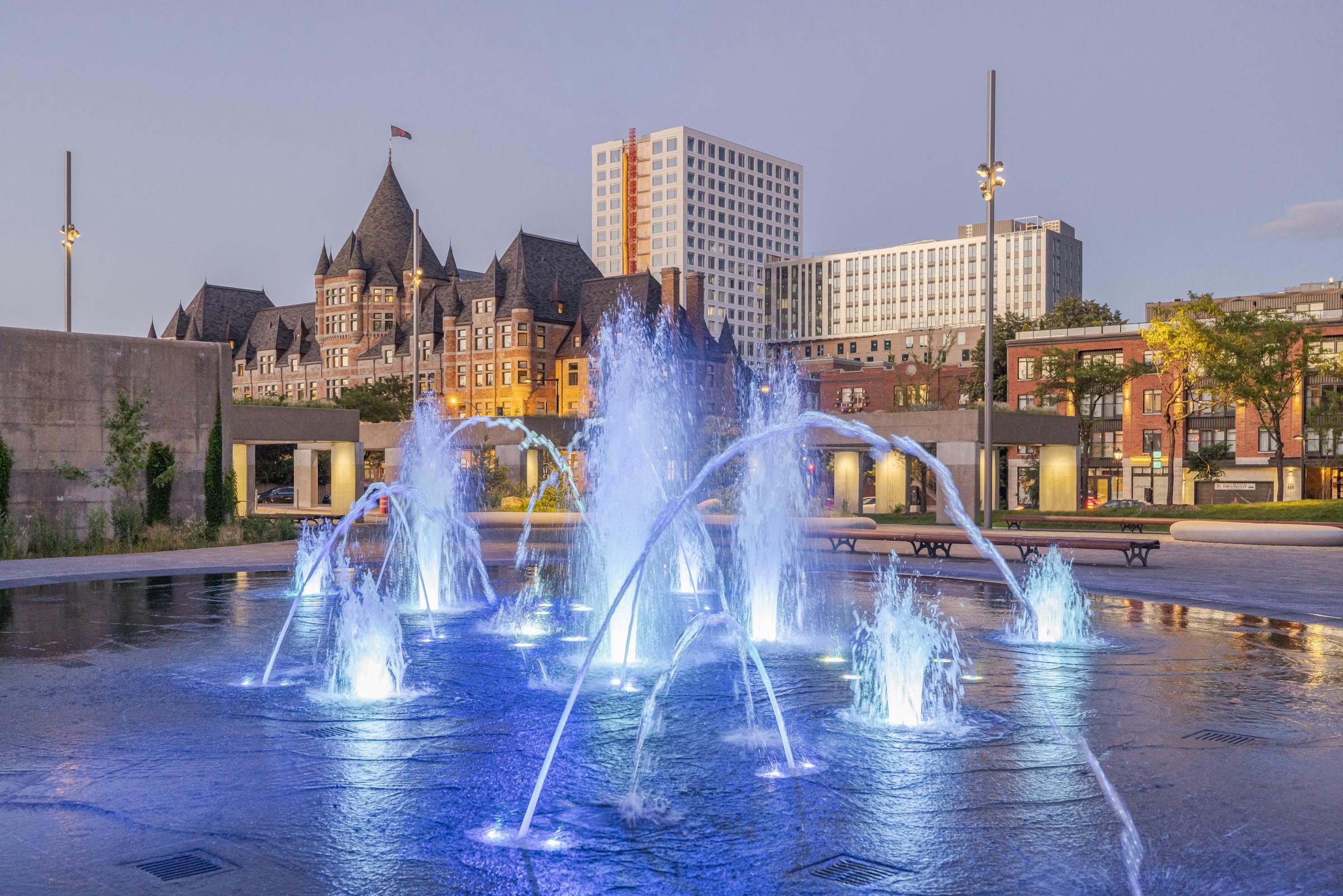 Photographie d'une fontaine retro éclairé en bleu avec des bâtiments architecturaux et patrimoniaux en arrière plan.