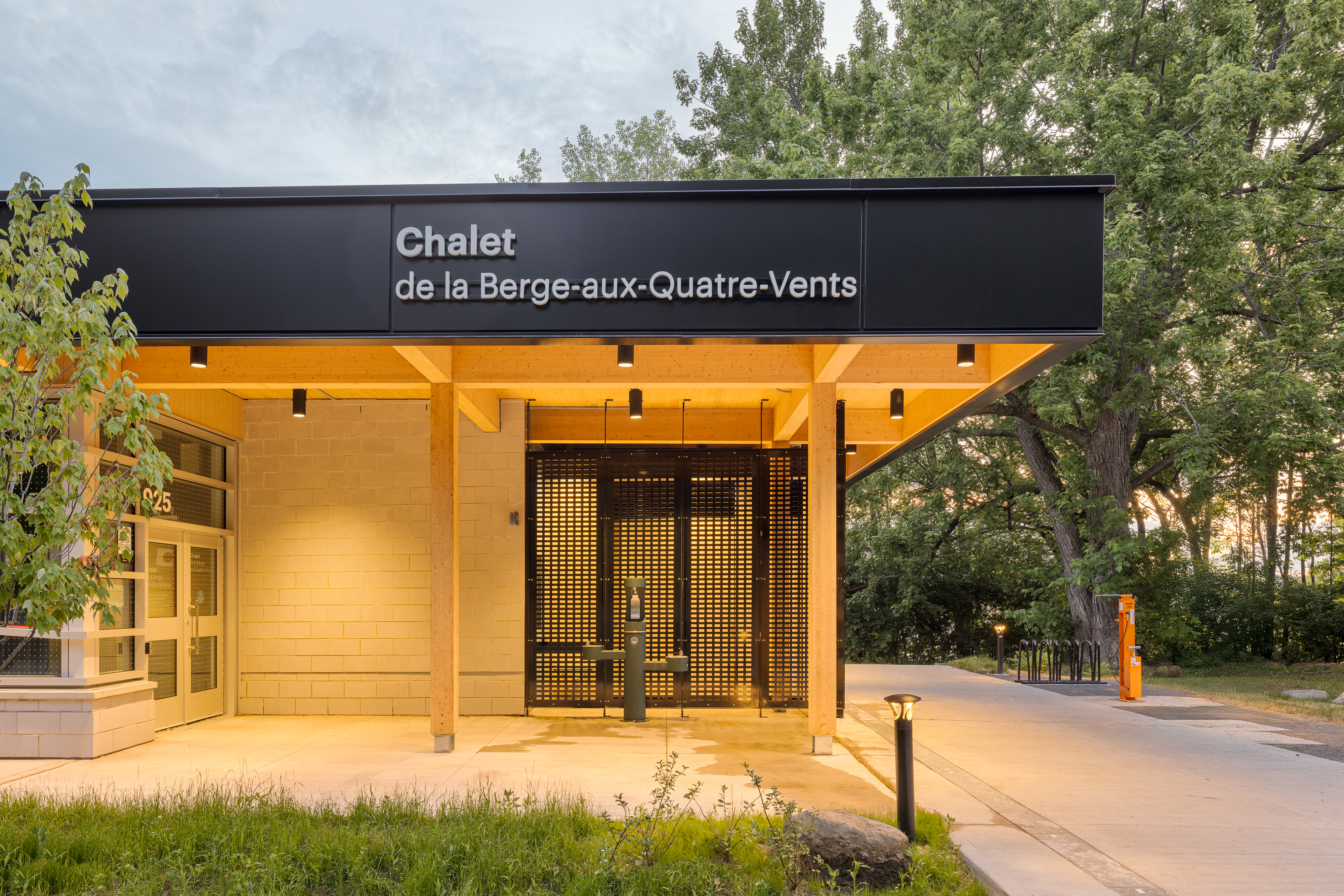 Photographie de l'entrée du chalet à l'heure dorée, avec l'enseigne Chalet de la Berge-aux-Quatre-Vents et l'éclairage architectural intégré à la structure de bois.