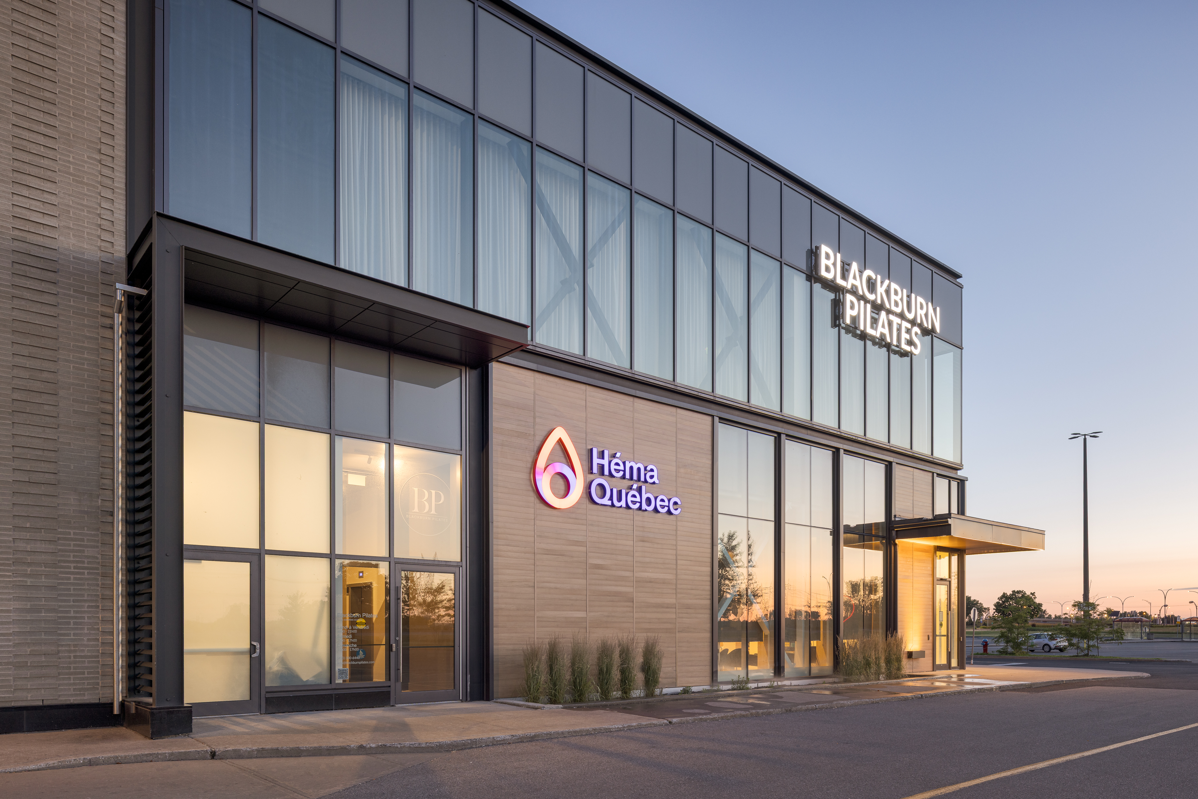 Photographie d'architecture commerciale du centre de dons Héma-Québec aux Promenades Saint-Bruno, à l'heure bleue, avec la façade de brique et l'enseigne voisine de Blackburn Pilates. ©TARMAC - Damien Ligiardi