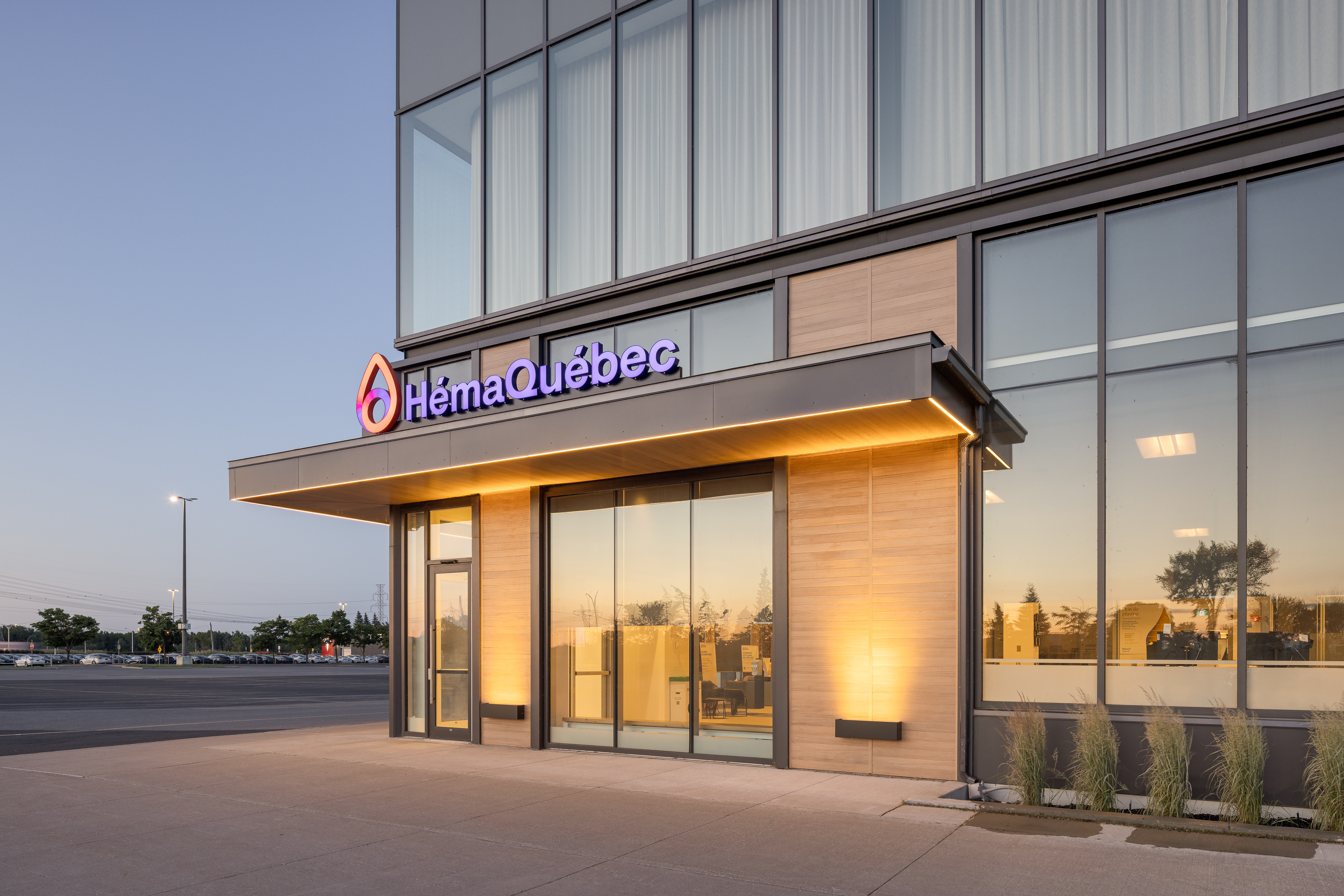 Photographie d'angle de l'entrée du centre Héma-Québec, mettant en valeur l'auvent, le mur-rideau vitré et le stationnement environnant au coucher du soleil. ©TARMAC - Damien Ligiardi