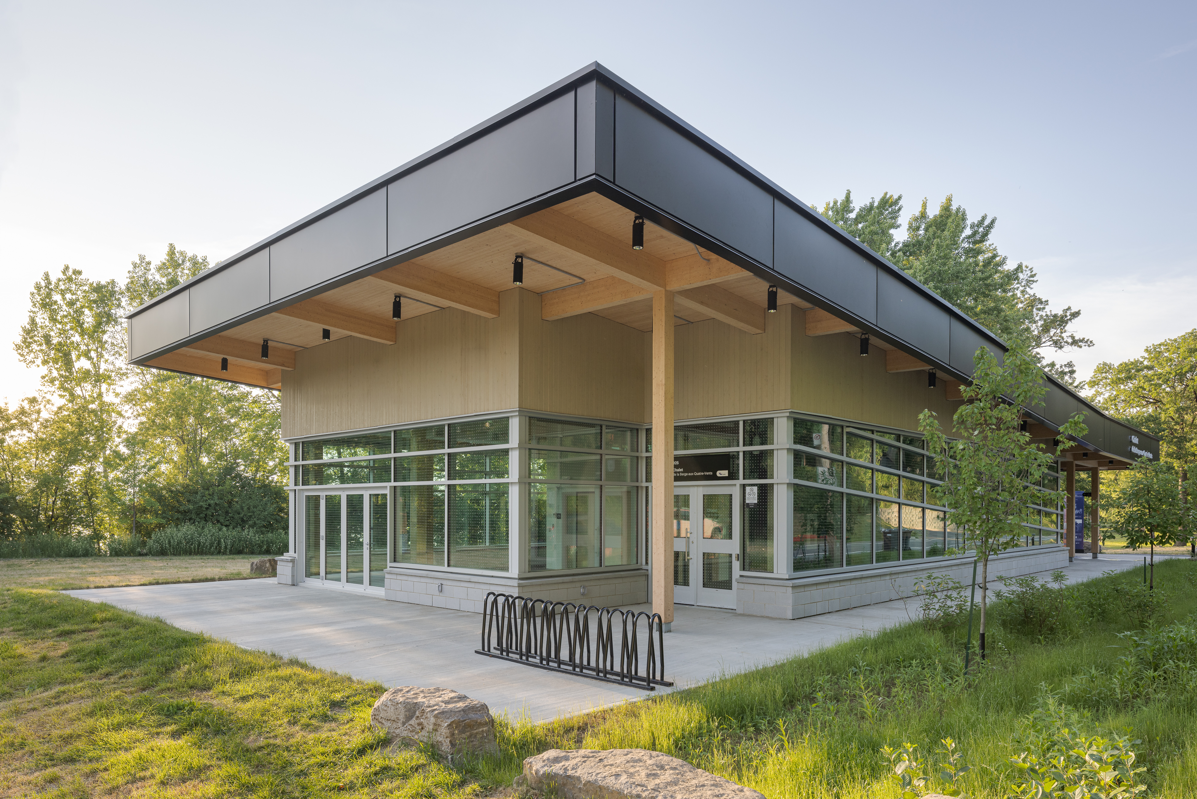 Photographie d'architecture de la façade vitrée du chalet, avec vue sur l'auvent de bois en porte-à-faux et l'aménagement paysager environnant.