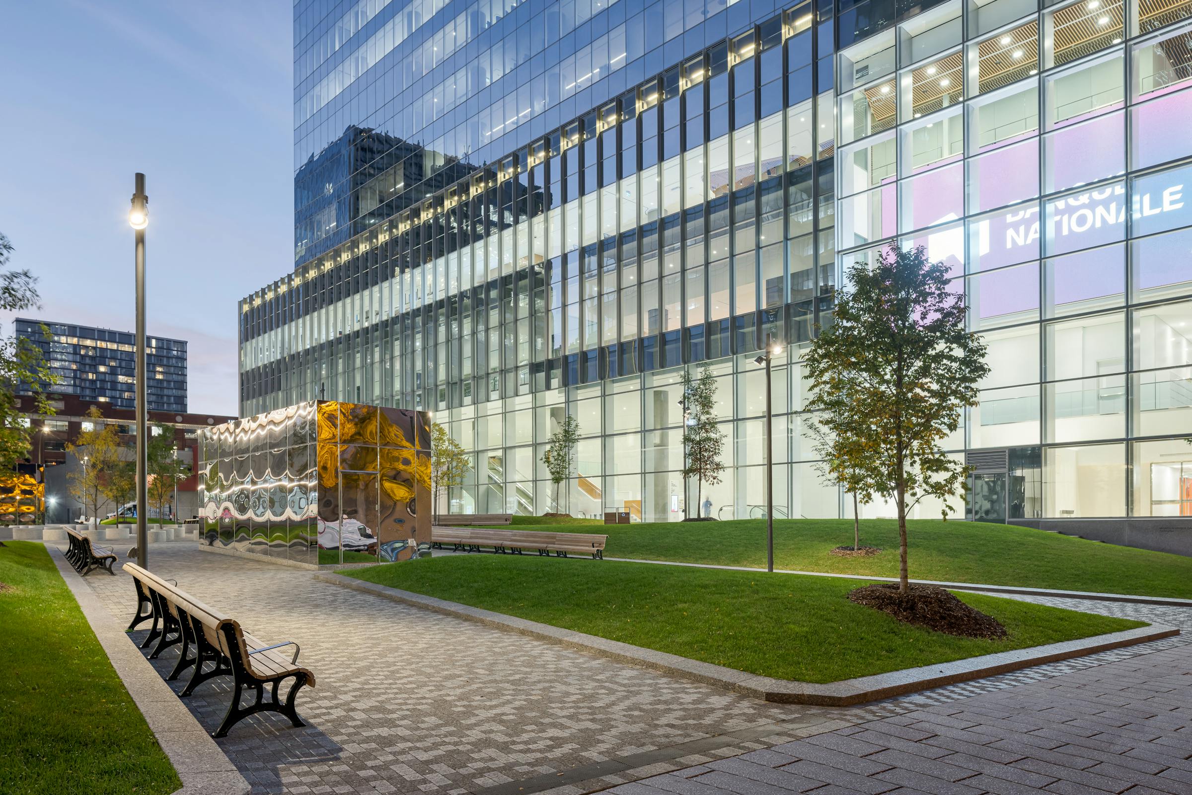 Vue oblique d’un espace pavé avec mobilier urbain et végétation, encadré par la tour de la Banque Nationale.