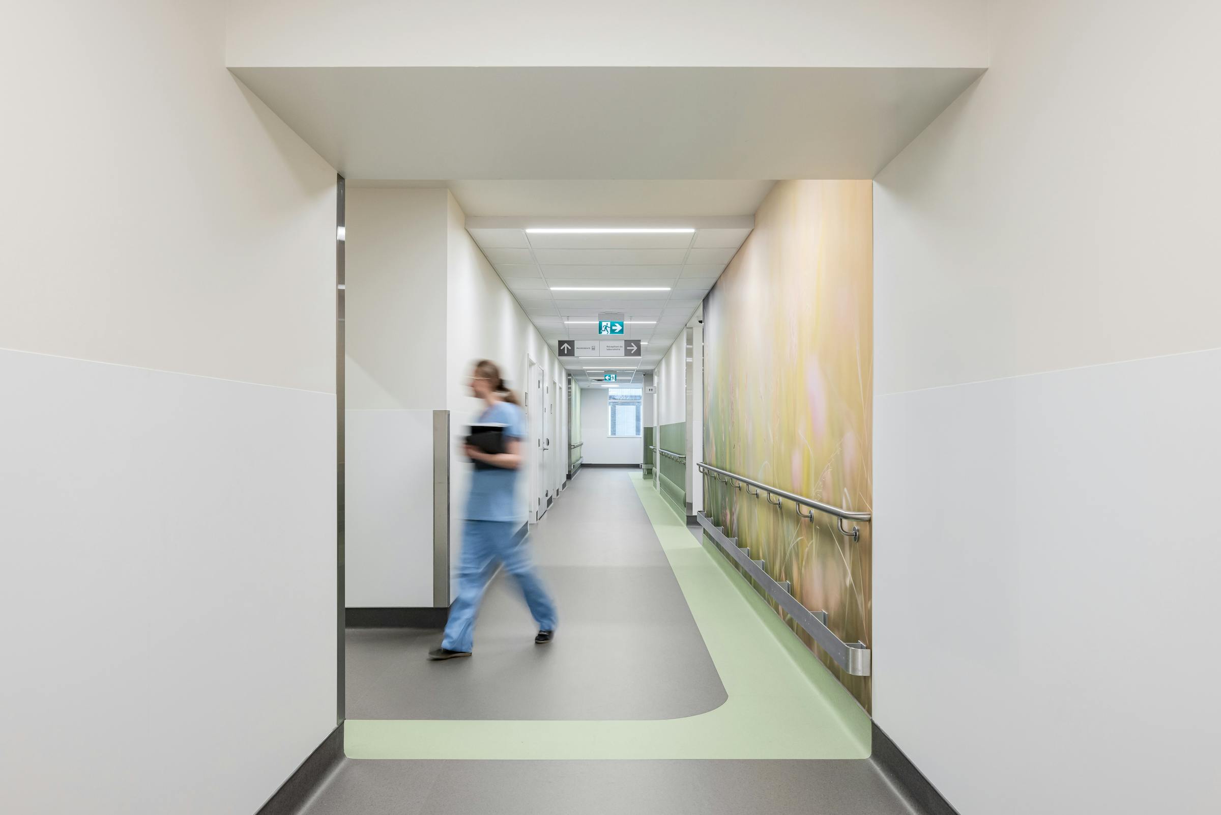 Photographie d’architecture d’un corridor d’hôpital lumineux avec fresque murale et personnel soignant en mouvement.