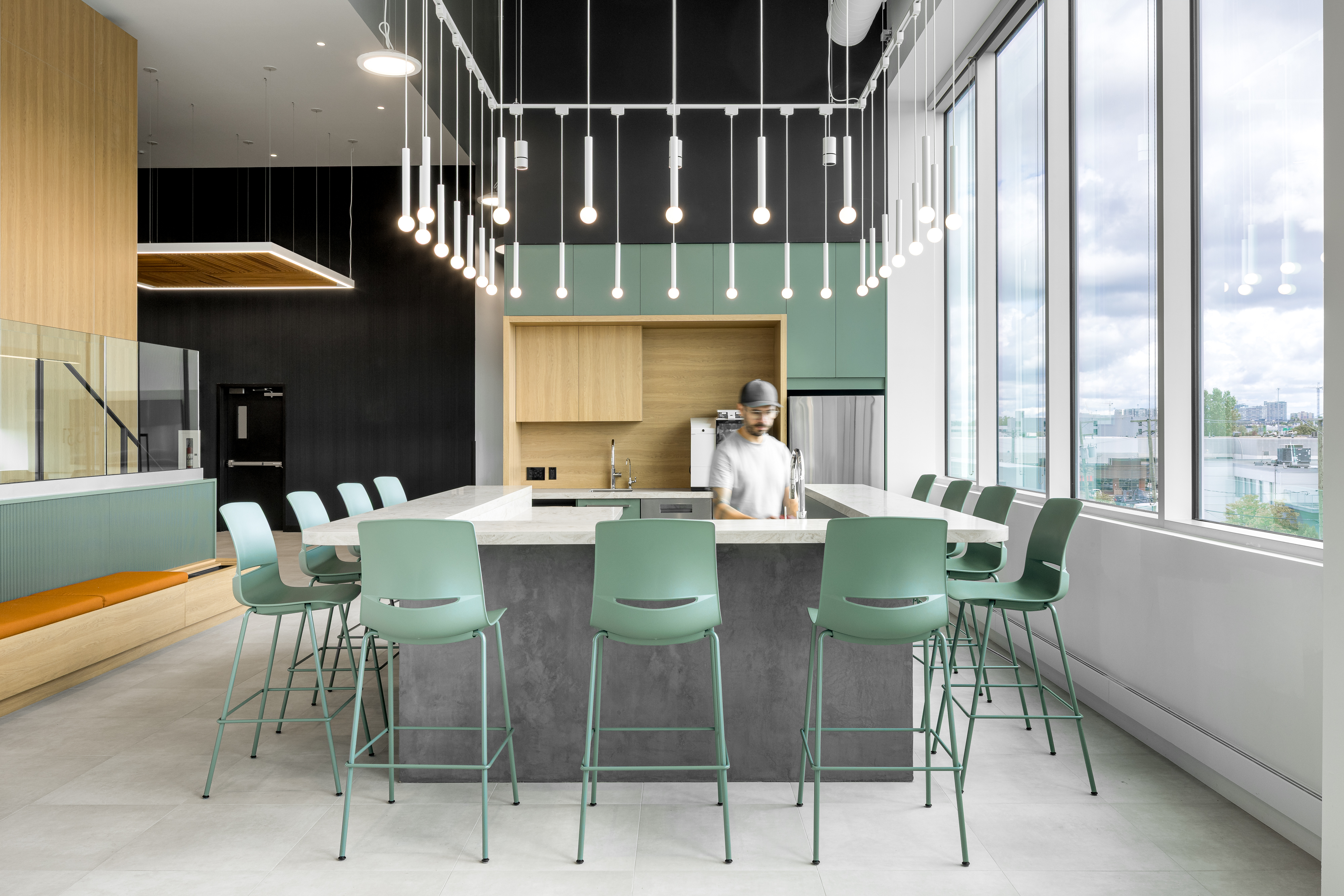 Photographie d’architecture d’une cafétéria de bureaux contemporaine avec îlot central, tabourets verts et luminaires suspendus.