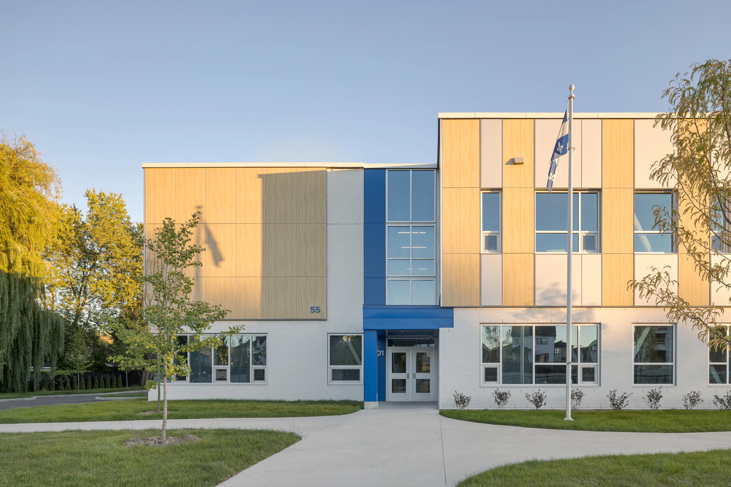 Façade principale de l'École des Saules, L'Assomption. Panneaux bois, aluminium et accents bleus au coucher du soleil. Photo : TARMAC - Damien Ligiardi.