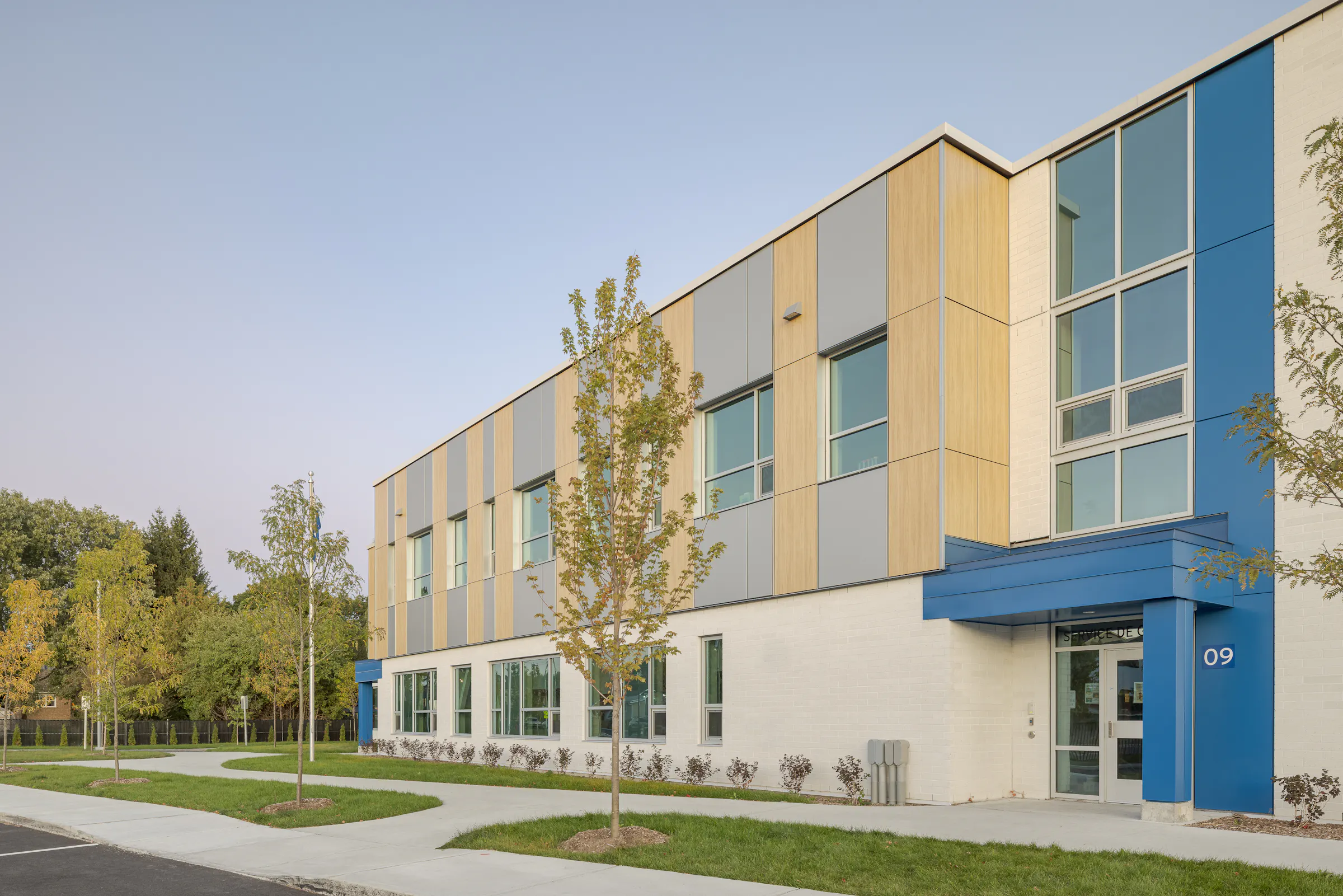 Entrée secondaire de l'École des Saules, L'Assomption. Façade bois et bleu, numérotation visible, ciel d'automne. Photo : TARMAC - Damien Ligiardi.
