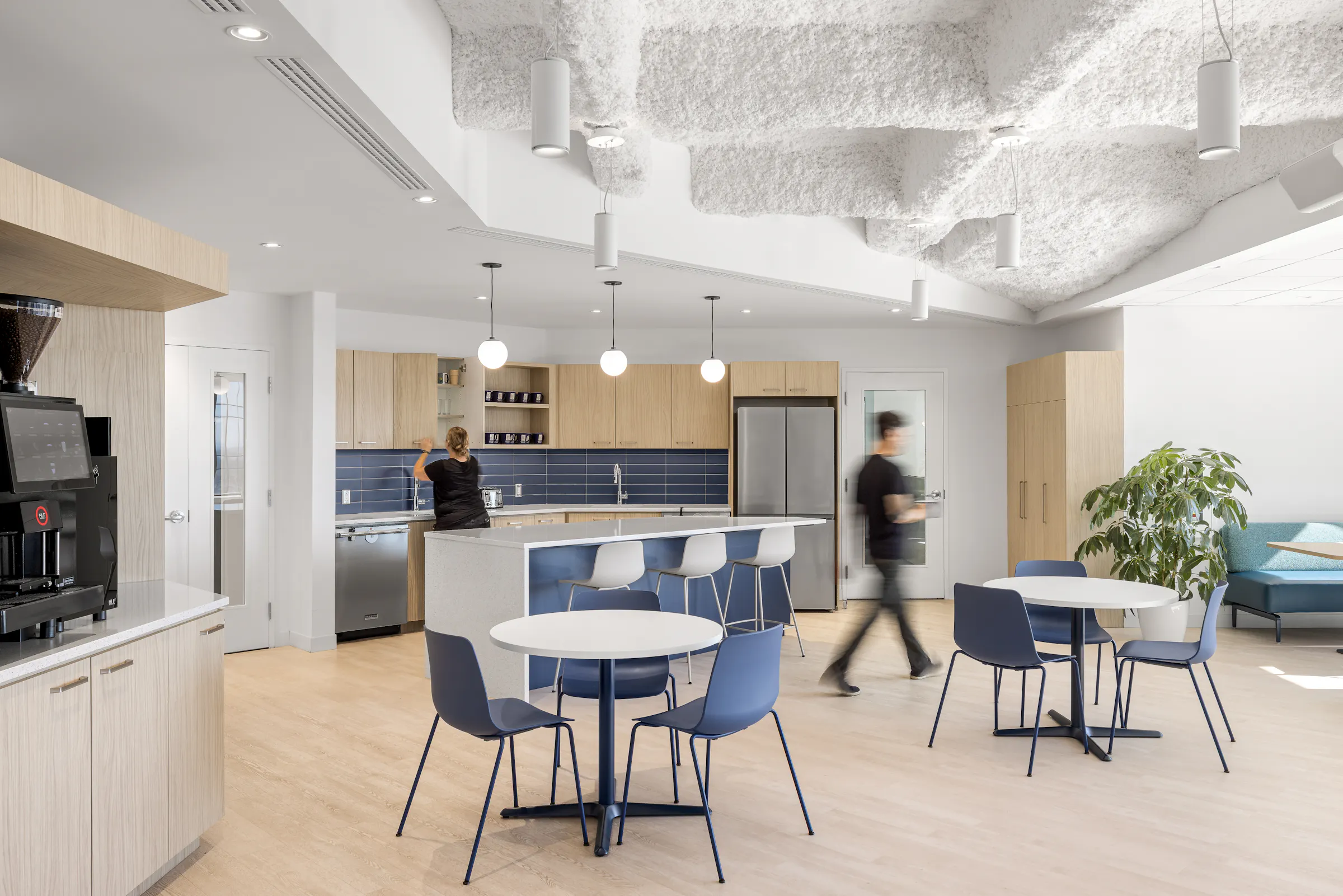 Espace cafétéria des bureaux d'ACI World à Montréal, avec plafond acoustique floconneux, tables blanches et chaises bleues, conçu par Conceptum International. Photo : TARMAC - Damien Ligiardi