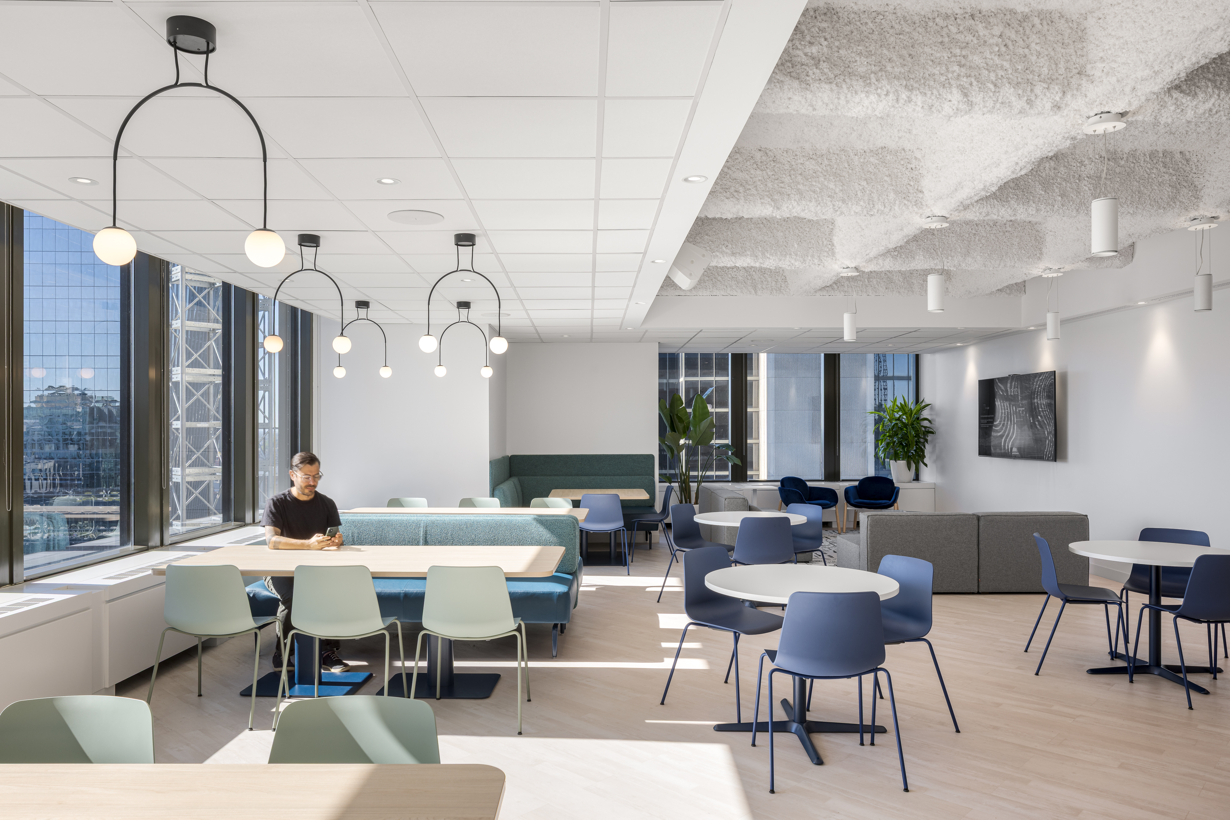 Cafétéria des bureaux d'ACI World à Montréal, avec plafond acoustique floconneux, luminaires design suspendus, tables rondes et chaises bleues et vertes, vue sur le centre-ville. Photo : TARMAC - Damien Ligiardi