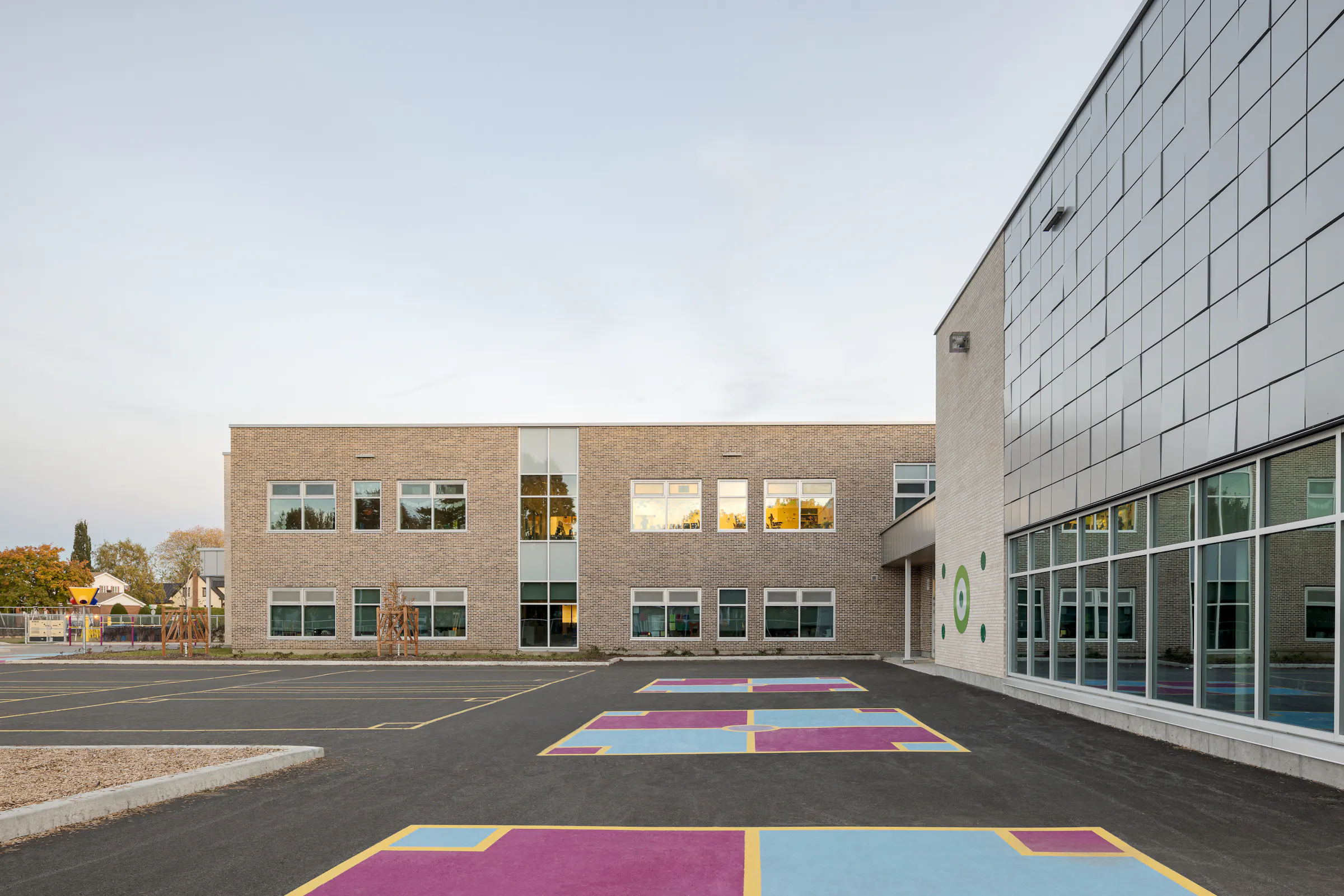Façade of the new wing at the end of the day, exterior lighting on, playground with coloured markings on the ground. Photo: TARMAC - Damien Ligiardi