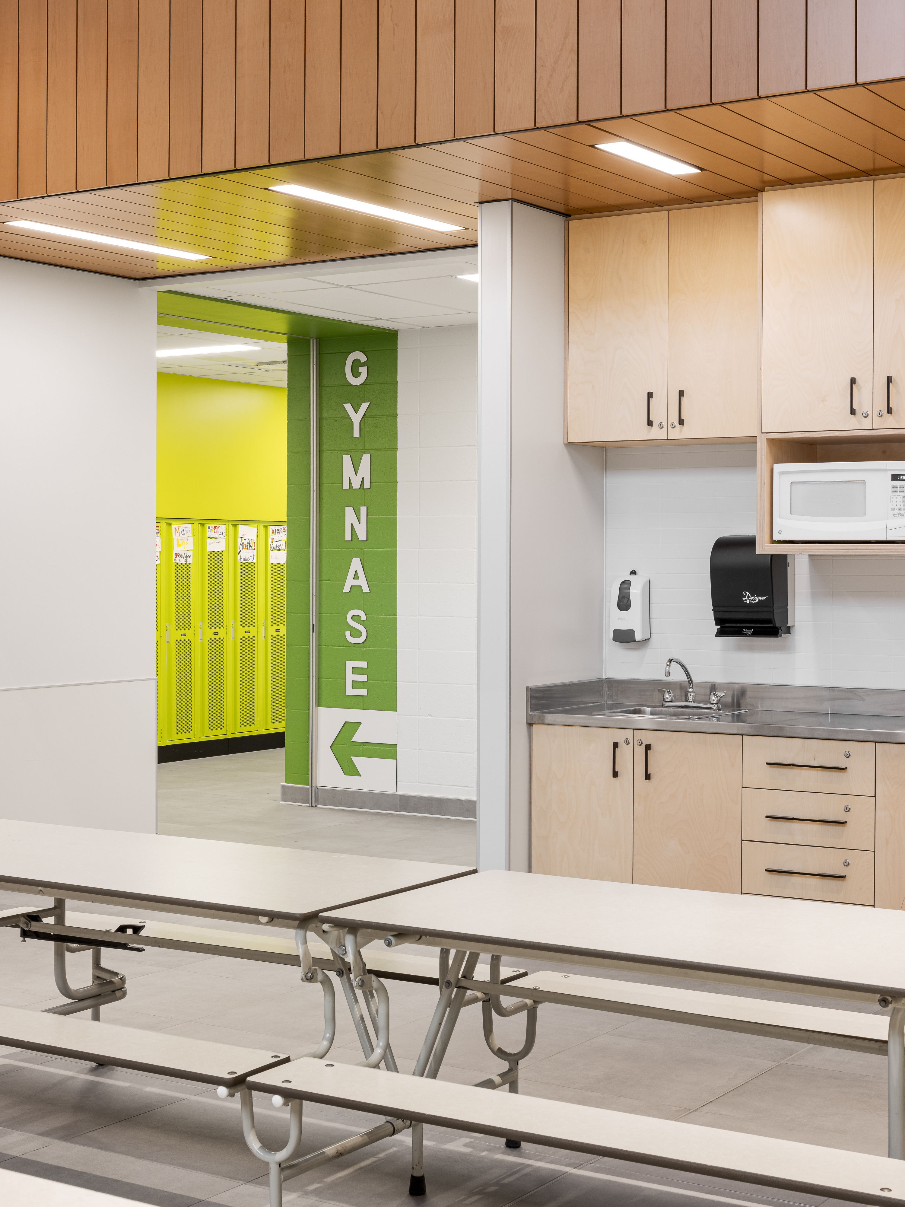 Cafeteria area with green directional signage to the gymnasium, wooden cabinets, and colorful accents. Photo: TARMAC - Damien Ligiardi