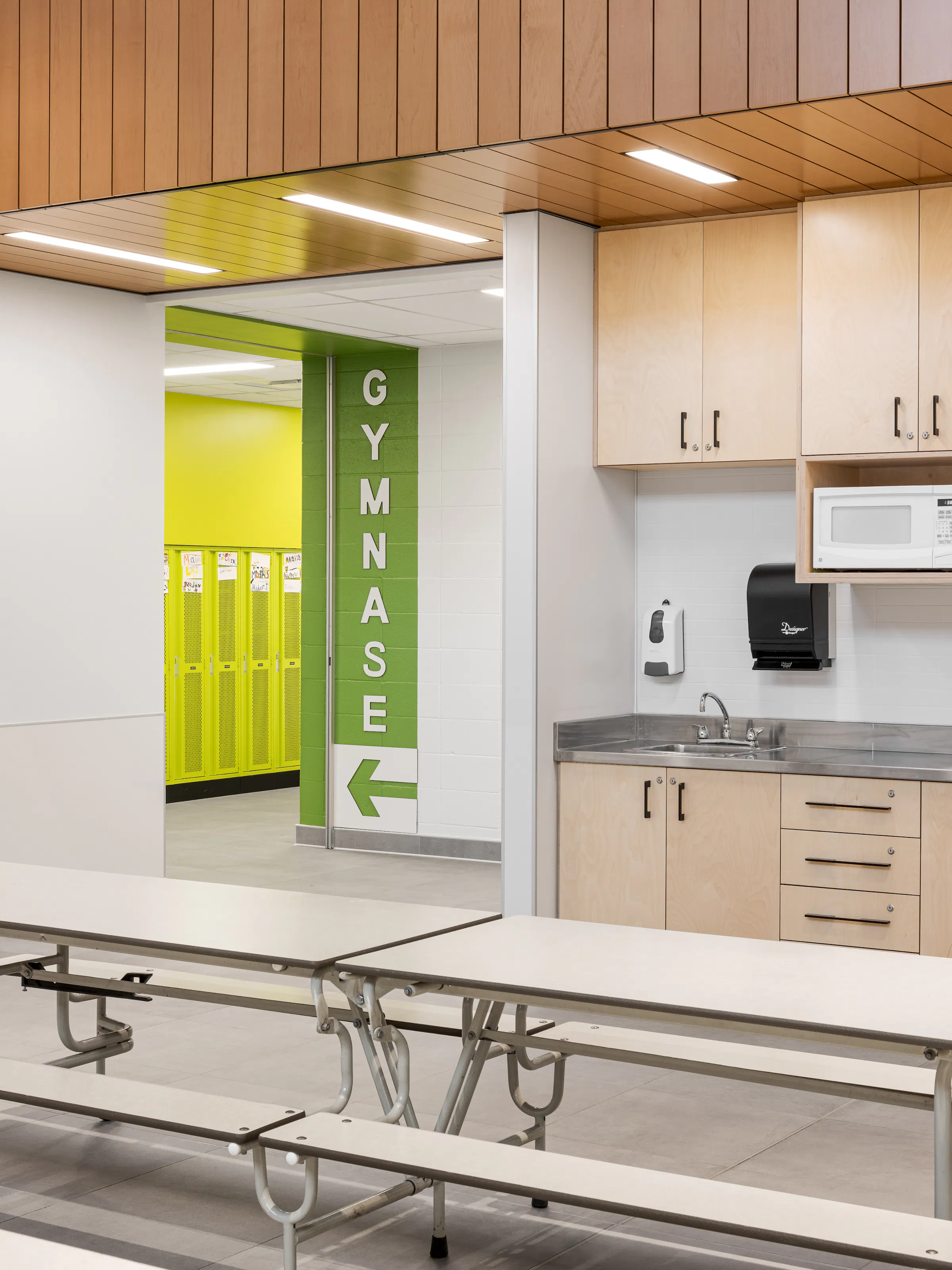 Cafeteria area with green directional signage to the gymnasium, wooden cabinets, and colorful accents. Photo: TARMAC - Damien Ligiardi
