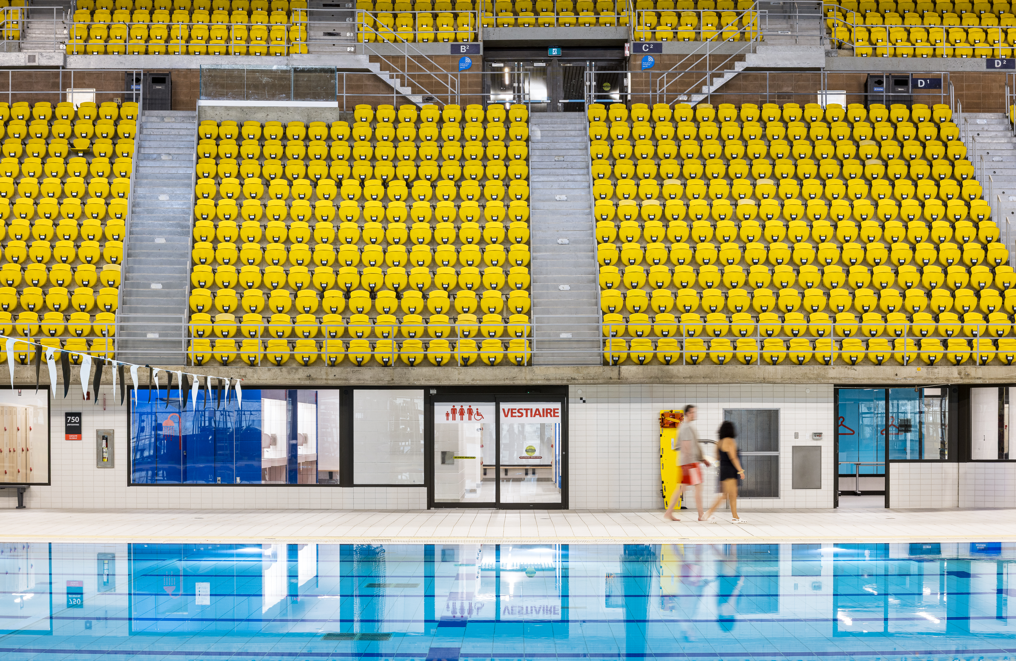 Photograph of an institutional building, yellow bleachers of the swimming pool with changing rooms in the background.