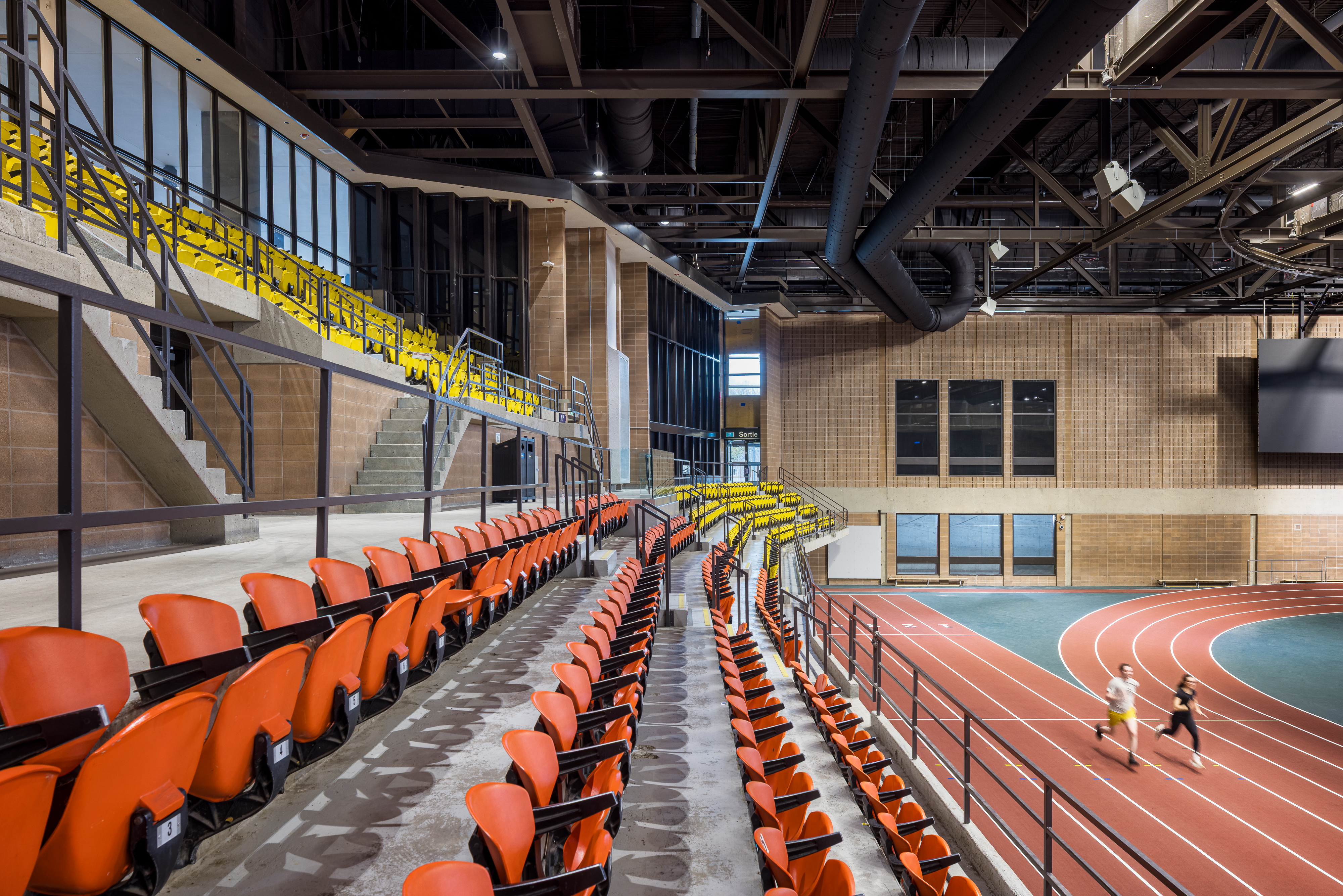 Photograph of sports architecture, yellow and orange bleachers overlooking the athletics track and tennis courts.