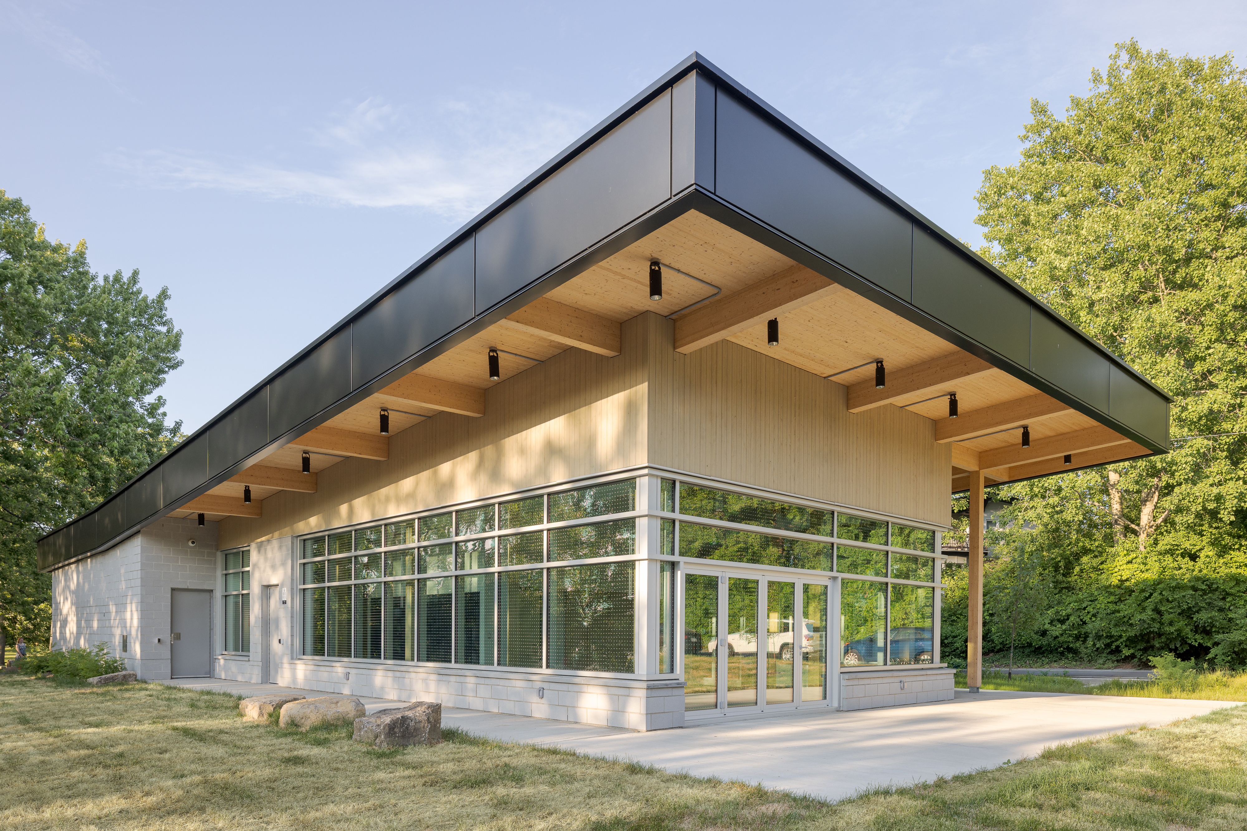 Photographie extérieure du chalet d'accueil de la berge aux Quatre-Vents à Laval, mettant en valeur le grand porte-à-faux du toit et la structure en bois intégrée au paysage.