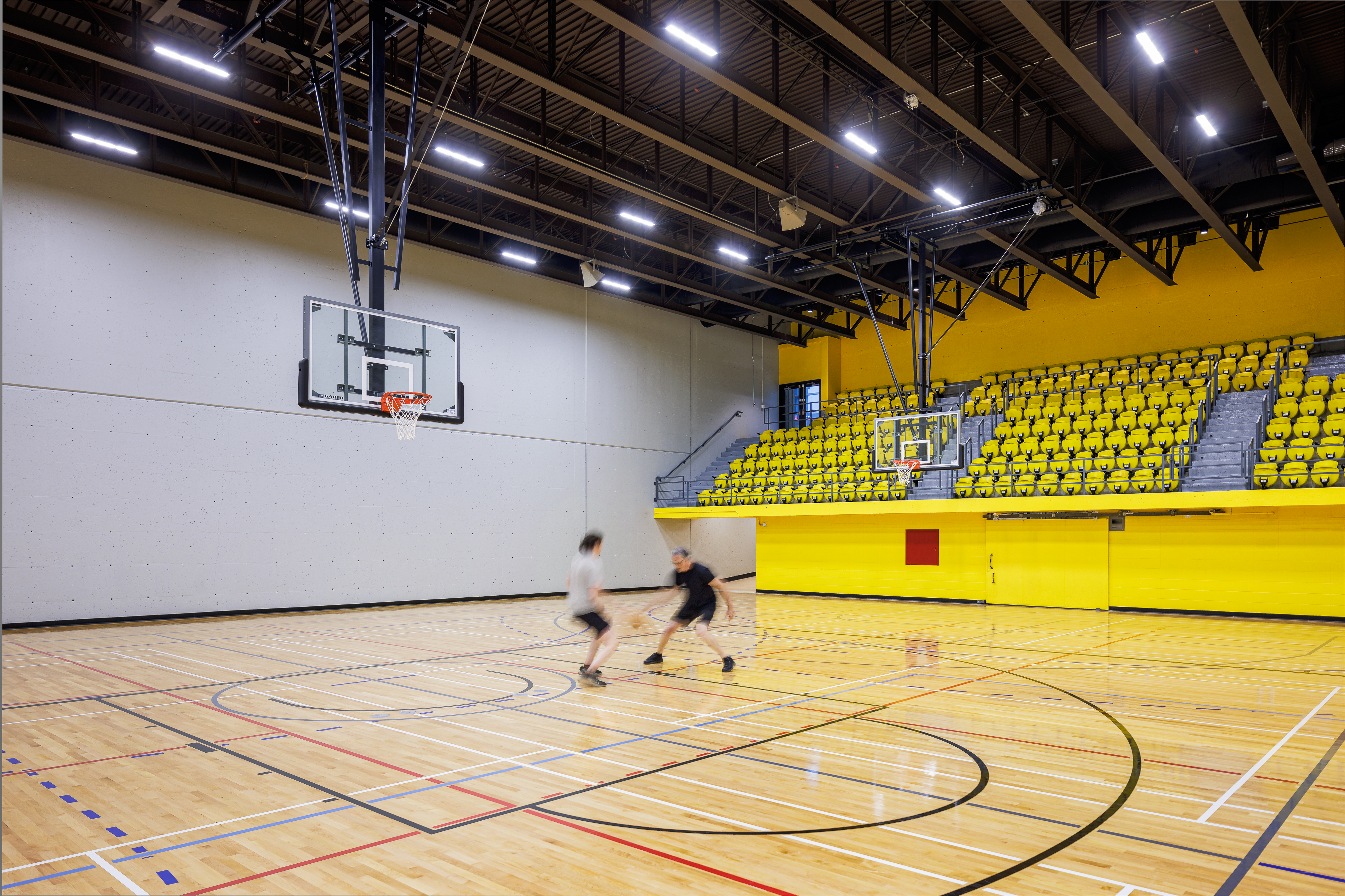 Photograph of sports architecture, basketball game in the renovated gymnasium, hardwood floor and yellow bleachers.
