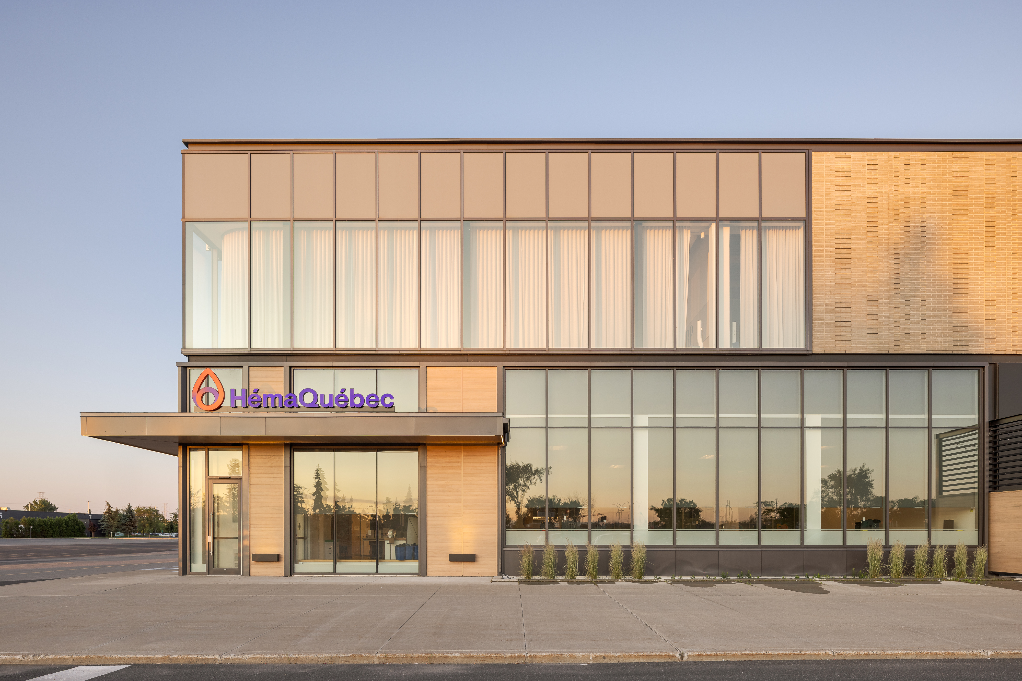 Photographie grand angle de la façade du centre Héma-Québec, montrant le mur-rideau vitré à l'étage et la signalétique au rez-de-chaussée au crépuscule. ©TARMAC - Damien Ligiardi