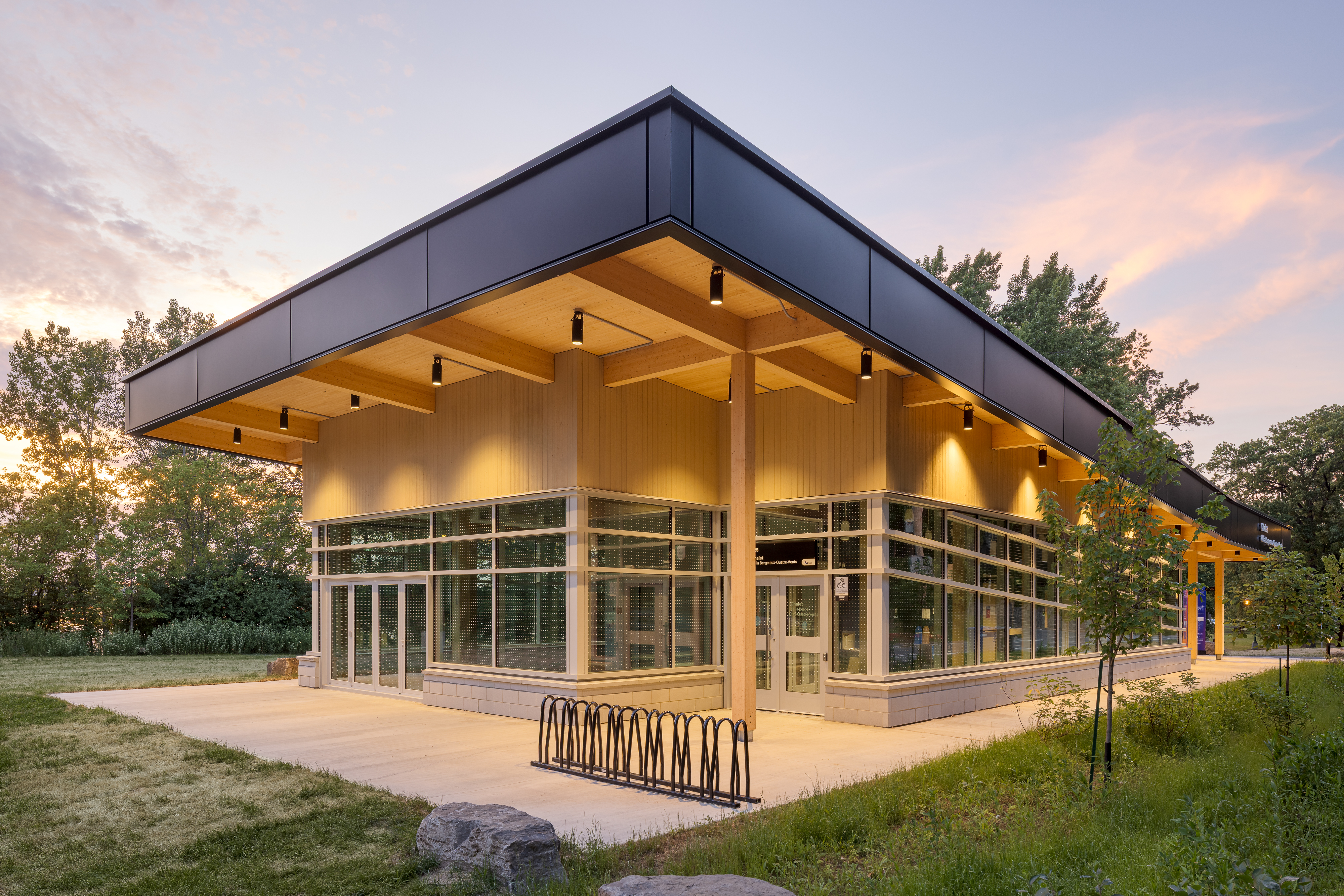 Photographie d'ambiance du chalet au coucher du soleil, avec la façade vitrée illuminée et le support à vélos à l'avant-plan.