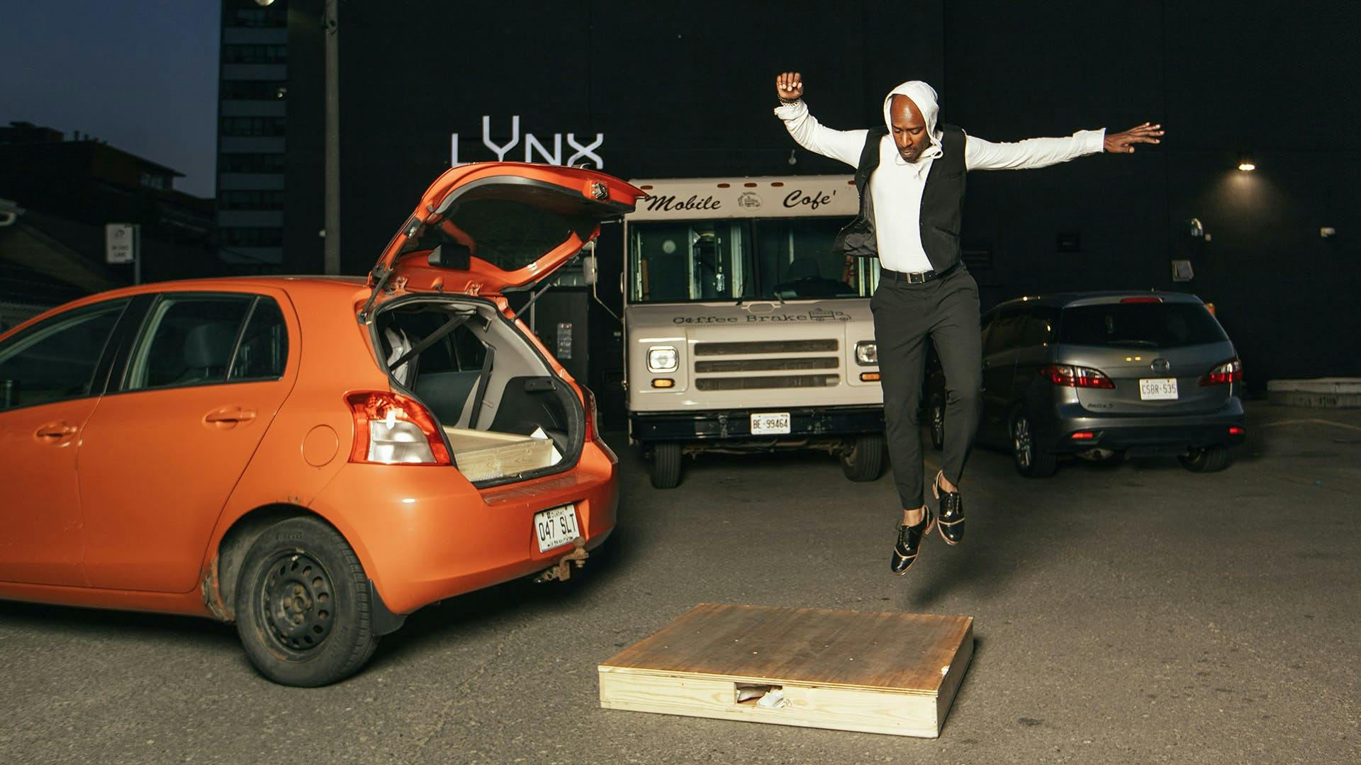 A person jumps next to a car with its boot open. 
