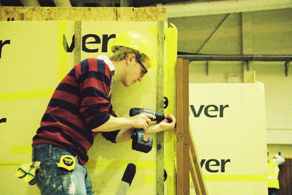 Tom drilling into the timber frame of the Nottingham H.O.U.S.E, wearing a hard hat and sporting badly dyed hair