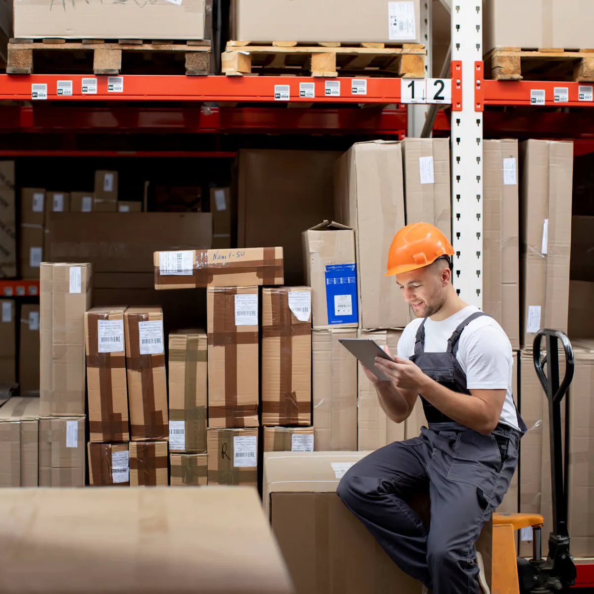Lagerarbeiter mit Schutzhelm und Hubwagen in einem Industrielager zwischen Kartons und Paletten – Intralogistik und Warehousing als Outsourcing-Lösung für die Lebensmittelindustrie.