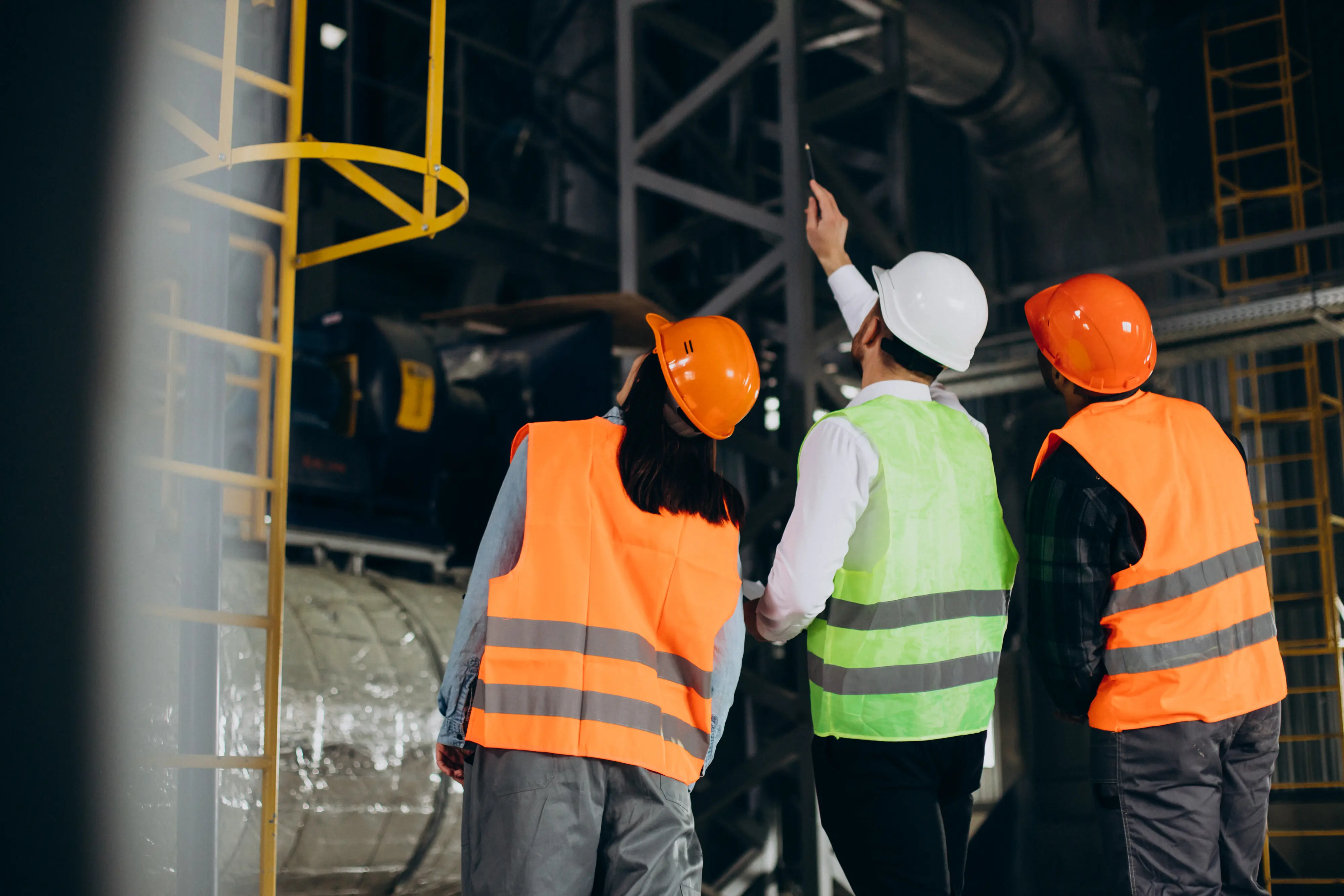  Drei Industriemitarbeiter mit Schutzhelmen und Warnwesten bei der Einsatzbesprechung in einer Industrieanlage – qualifizierte Sicherungsposten für Arbeiten in engen Räumen und Gefahrenbereichen nach DGUV 113-004.