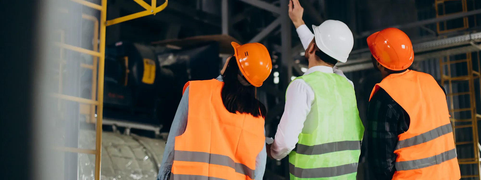 Drei Industriemitarbeiter mit Schutzhelmen und Warnwesten bei der Einsatzbesprechung in einer Industrieanlage – qualifizierte Sicherungsposten für Arbeiten in engen Räumen und Gefahrenbereichen nach DGUV 113-004.