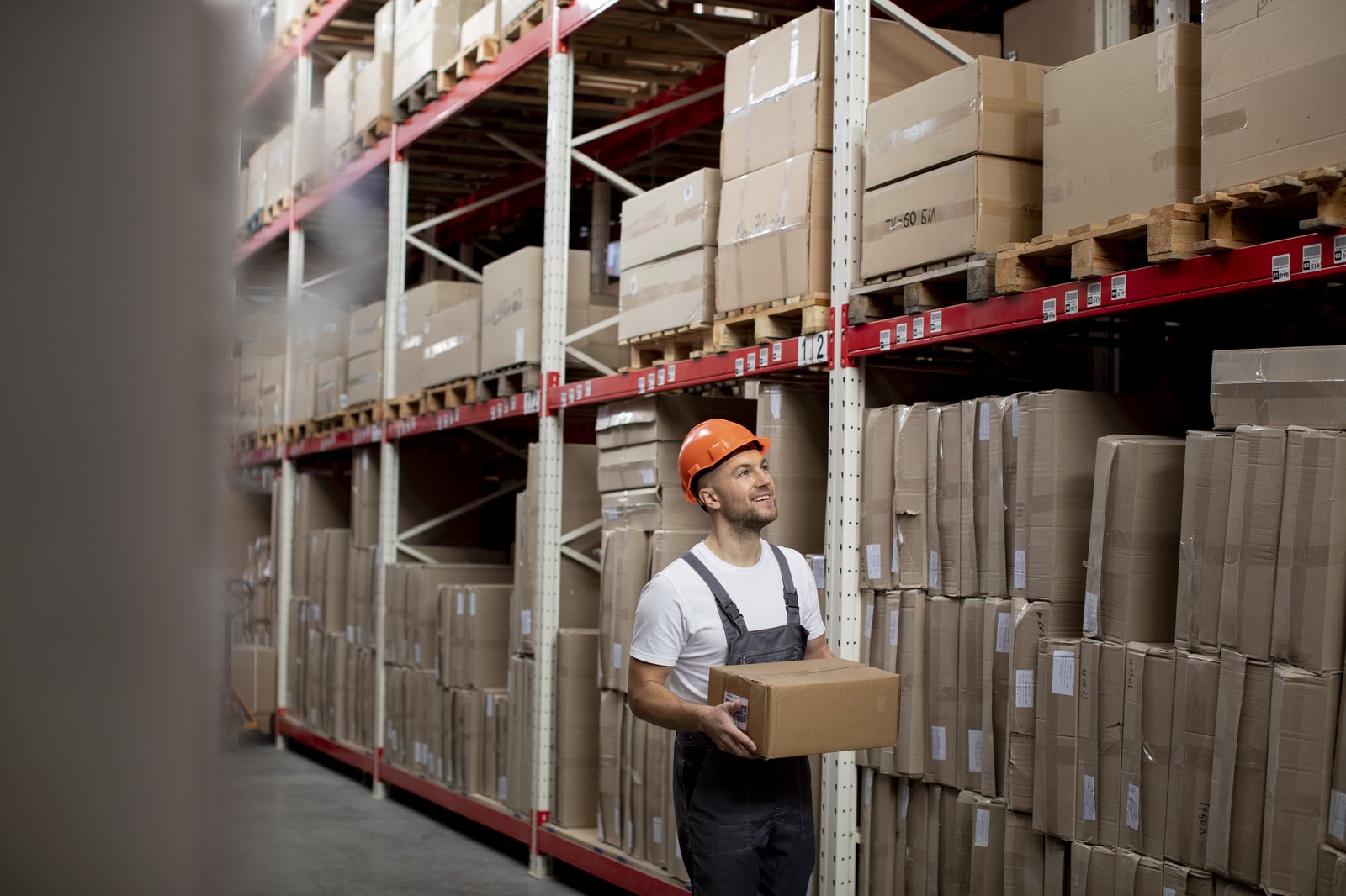 Lagerarbeiter mit Schutzhelm trägt Karton im Hochregallager – Lagerlogistik Outsourcing durch Industriedienstleister
