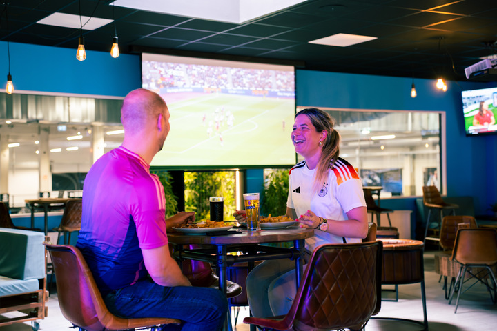 Zwei Gäste in Trikots sitzen am Tisch, essen und schauen Fußball