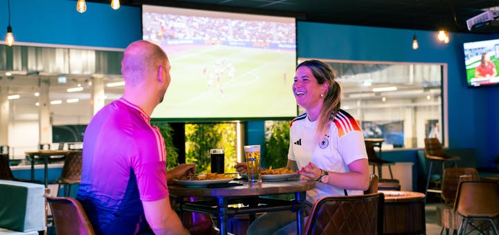 Zwei Gäste in Trikots sitzen am Tisch, essen und schauen Fußball