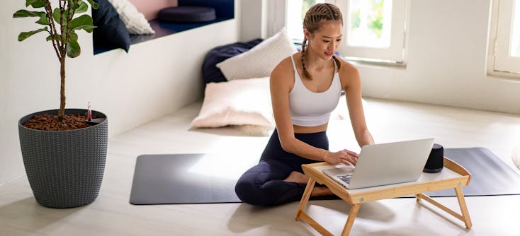 A fitness instructor sitting on a yoga mat reading a fitness blog on their laptop.
